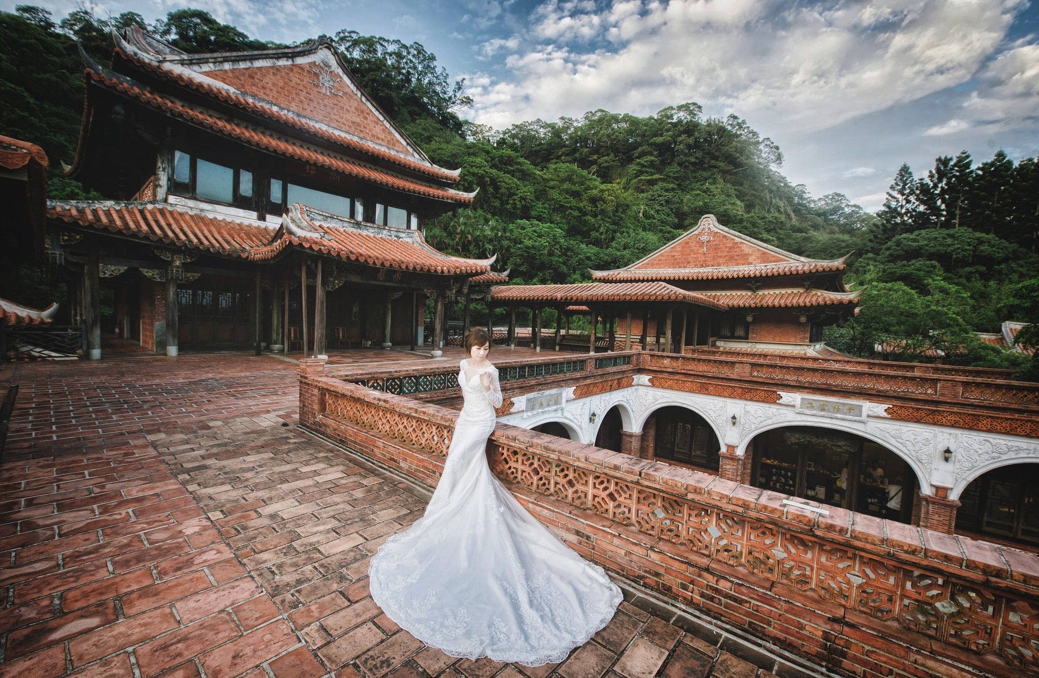 廟宇婚紗 壁紙 — 一位身穿飄逸白色婚紗的新娘，站在鋪有赤陶磚的陽台上，俯瞰著傳統東亞寺廟建築，這些建築坐落在鬱鬱蔥蔥的綠色山丘中，天空部分多雲。