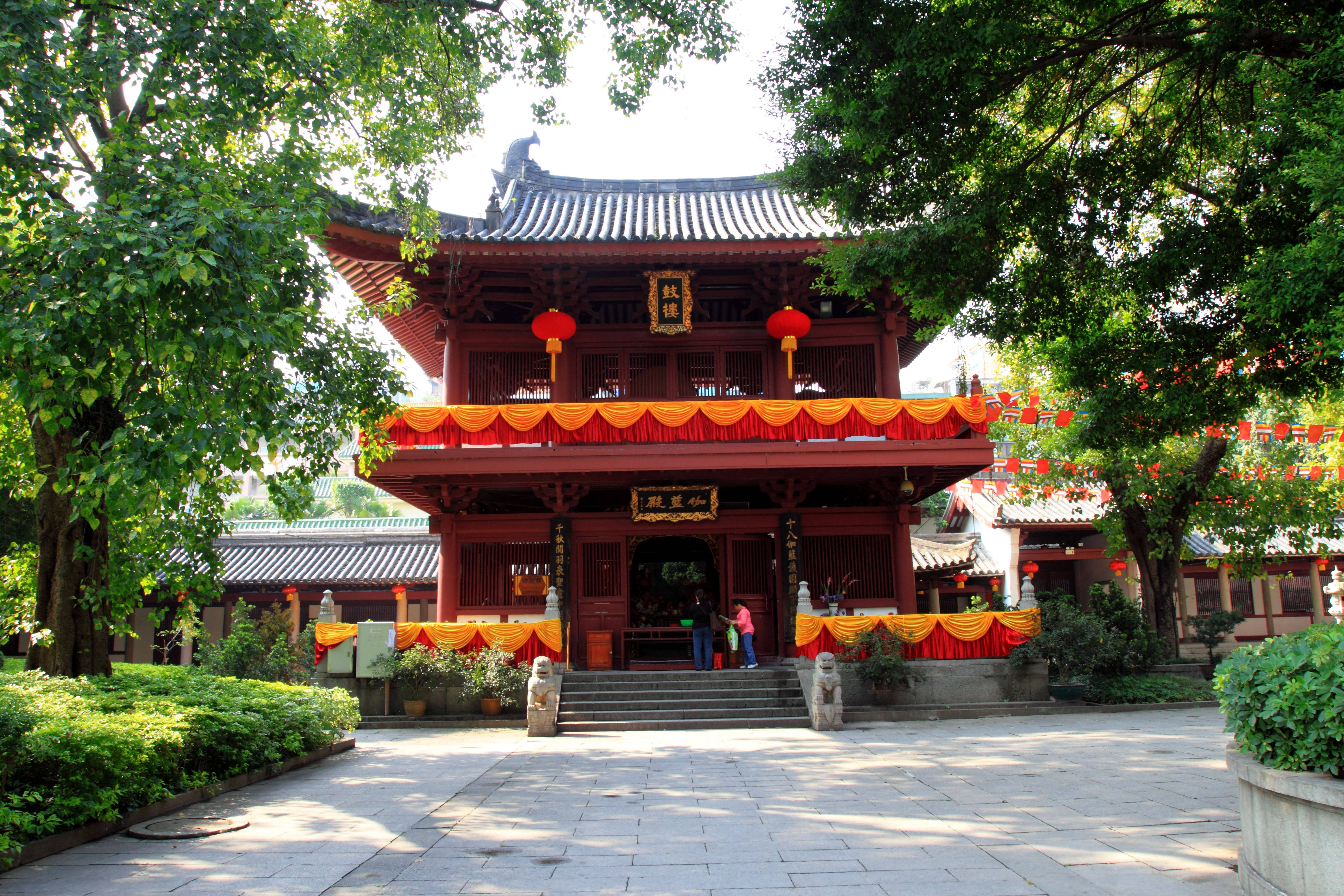 廣州光孝寺 壁紙 — 一座傳統的紅色木造寺廟，擁有華麗的屋頂和橙色窗簾，在晴朗的日子裡矗立在明亮的綠樹下。