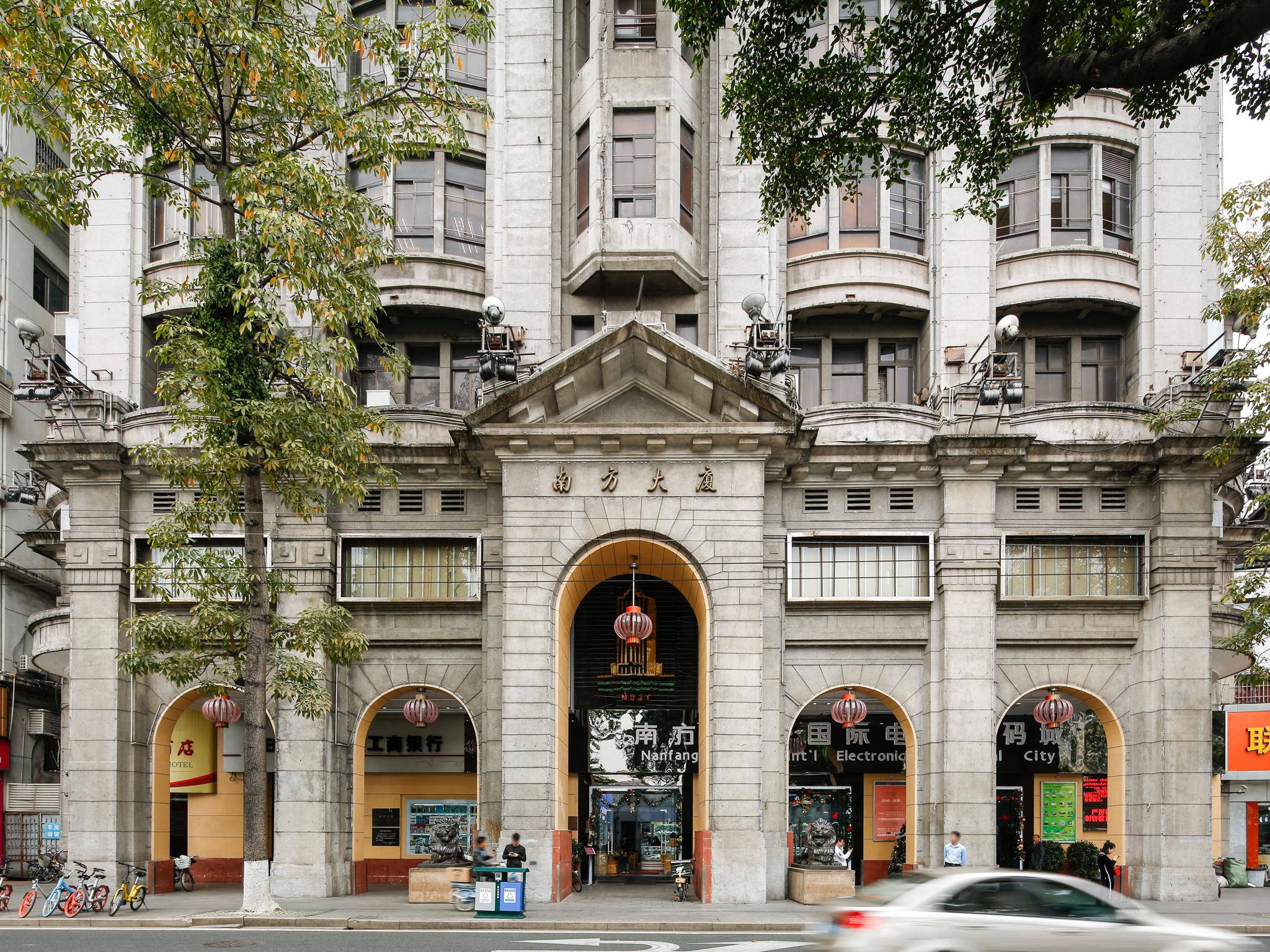 廣州南方大廈 壁紙 — 大型石面建築，設有拱形入口和中文字，周圍環繞著茂密的綠樹和一輛經過的汽車。