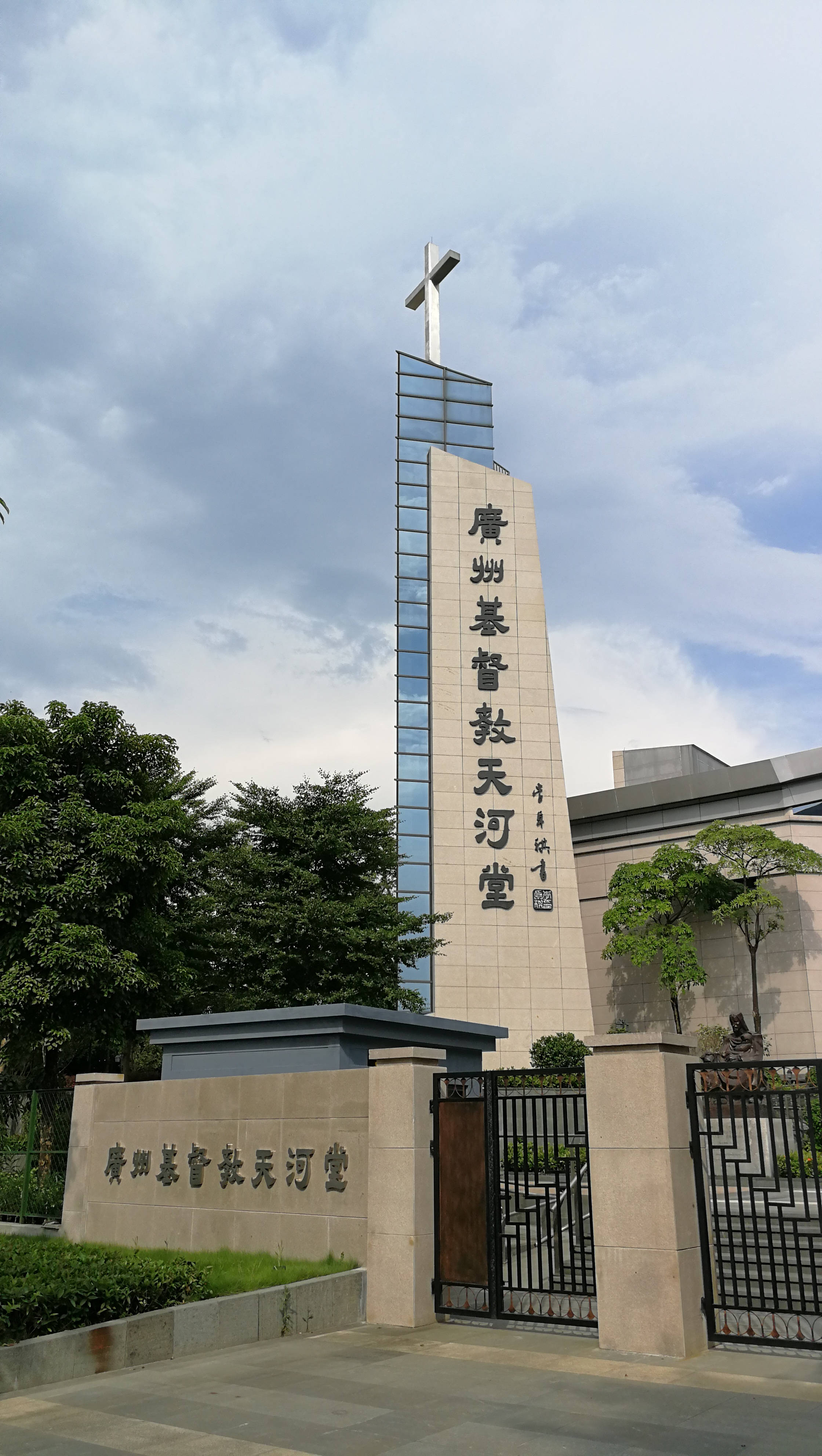 廣州天河教堂 壁紙 — 一座現代教堂，擁有高聳的玻璃和石頭塔樓，頂端有十字架，矗立在多雲的天空下，茂密的綠樹環繞著其有門的入口。