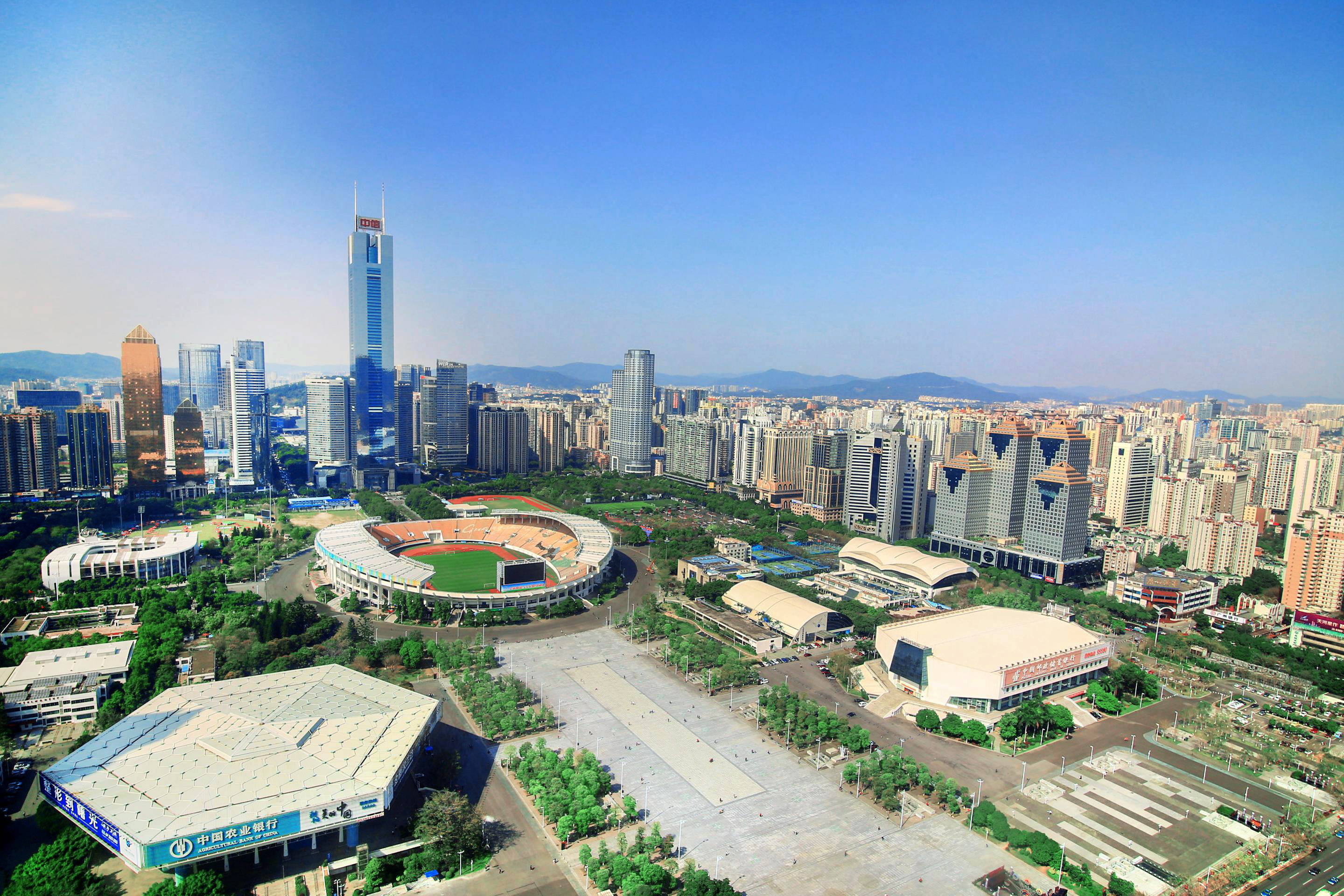 廣州天河體育中心 壁紙 — 現代城市景觀的鳥瞰圖，包括摩天大樓、一個大型體育館和綠地，在晴朗的藍天下。