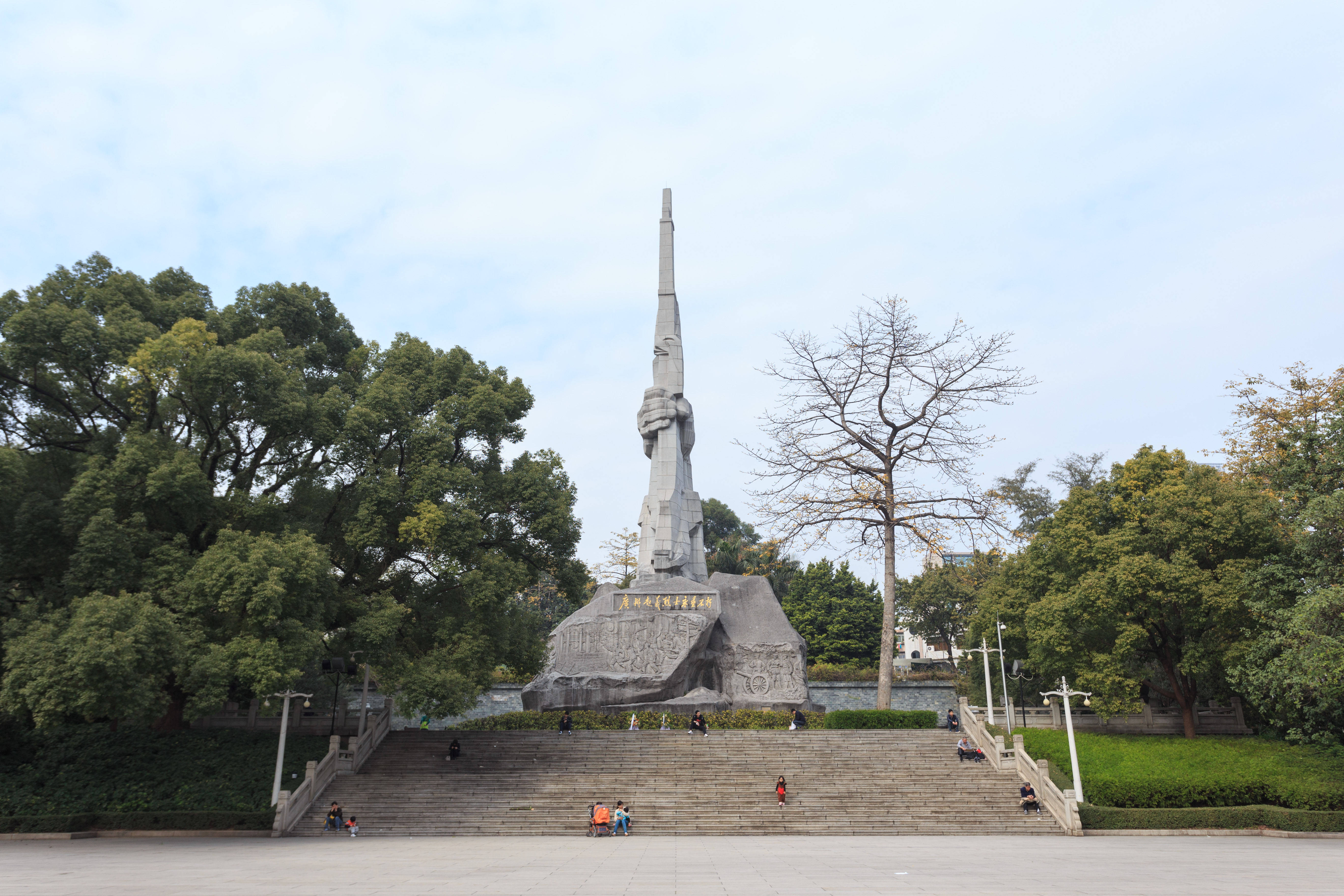 廣州烈士紀念園 壁紙 — 一座高聳的灰色紀念碑，頂部有緊握拳頭的雕塑，矗立在寬闊的石階上，周圍是茂密的綠樹，天空是淡藍色。