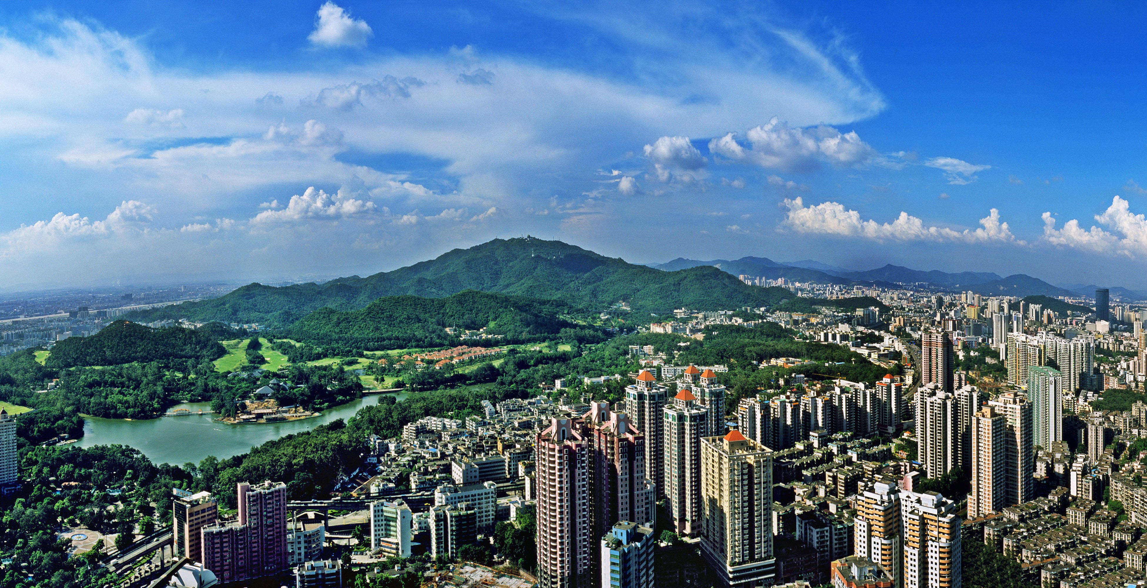廣州白云山 壁紙 — 俯瞰一座現代化城市，蜿蜒的河流和鬱鬱蔥蔥的綠色山丘環繞，明亮的藍天白雲點綴。