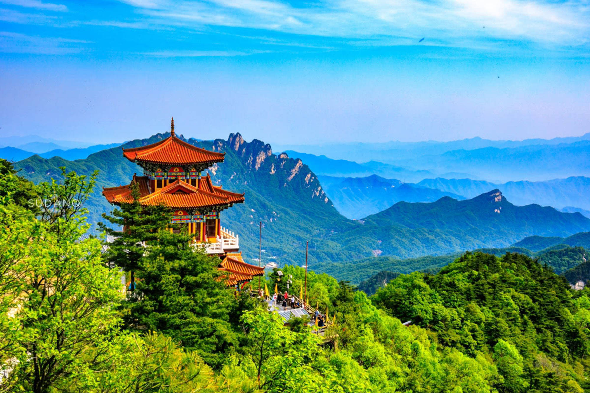 廣州白雲山 壁紙 — 一座傳統橙色屋頂的寺廟坐落在鬱鬱蔥蔥的綠色山頂上，俯瞰著晴朗天空下層疊的藍色山峰。