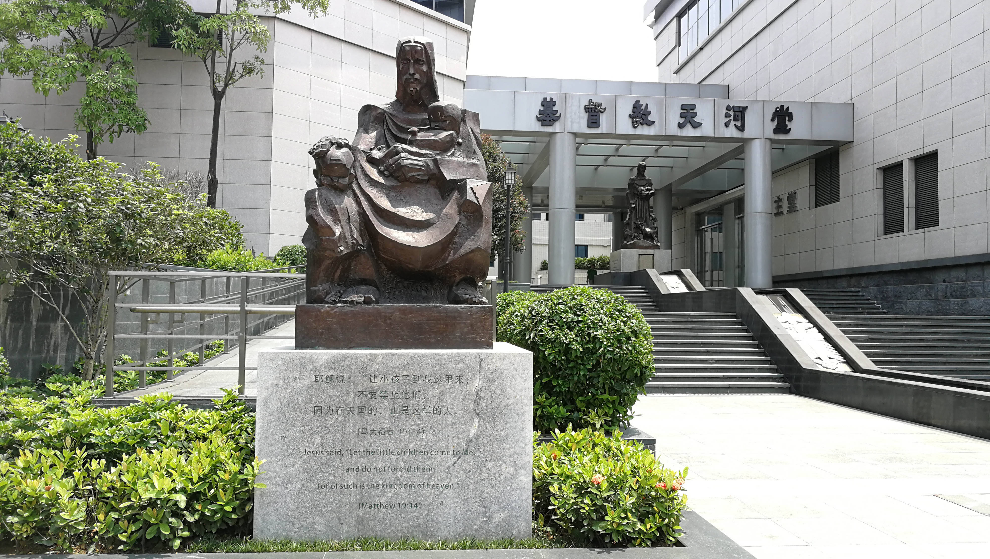 廣州耶穌愛孩子雕像 壁紙 — 一座青銅雕像，描繪一位坐著的人物抱著一個孩子，矗立在一棟現代建築入口前，周圍環繞著綠色灌木和石階。