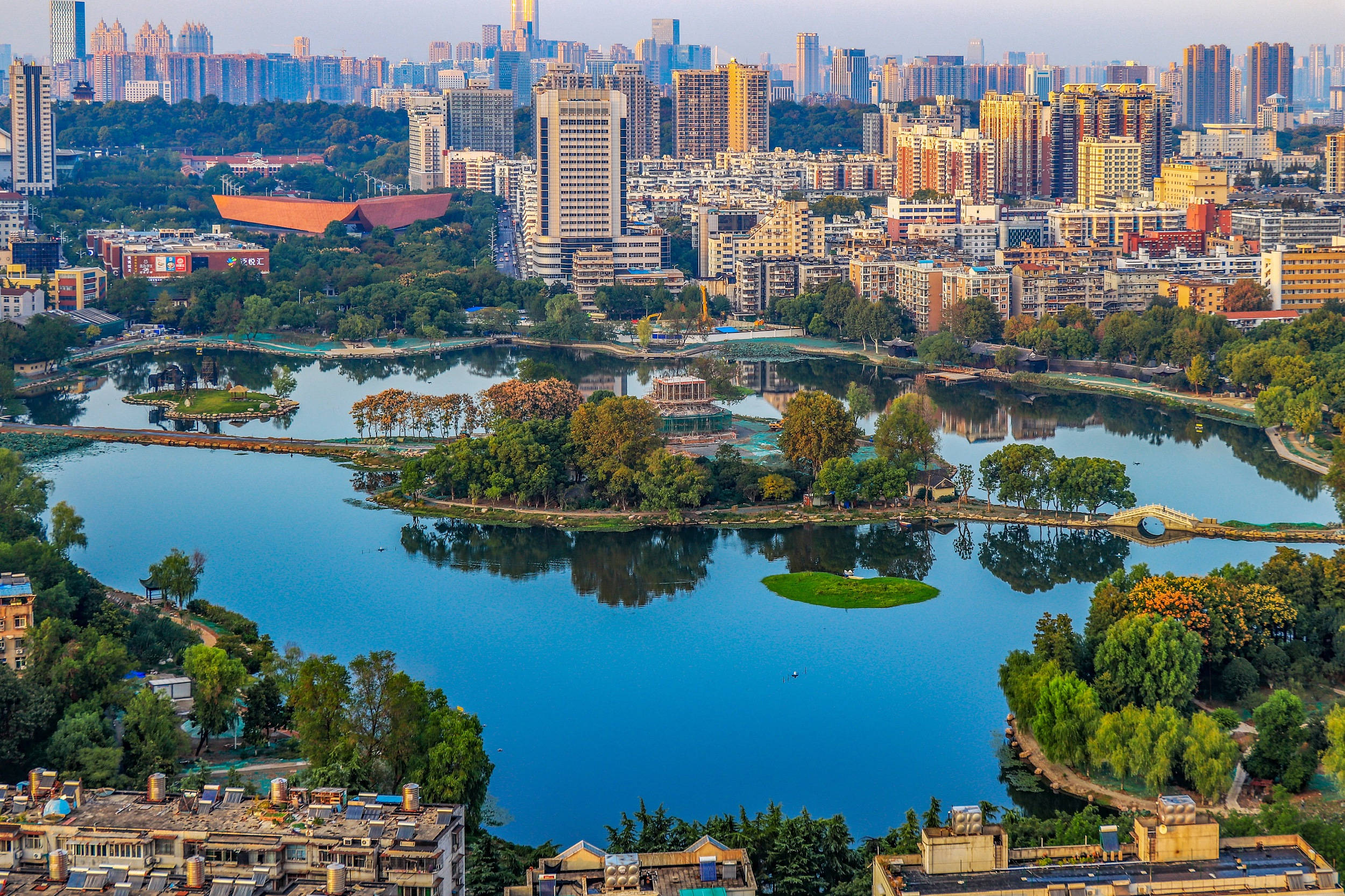 廣州荔灣湖公園美景 壁紙 — 從空中俯瞰，一個寧靜的城市湖泊被茂密的綠化和現代高層建築環繞，沐浴在柔和的金色時光中。