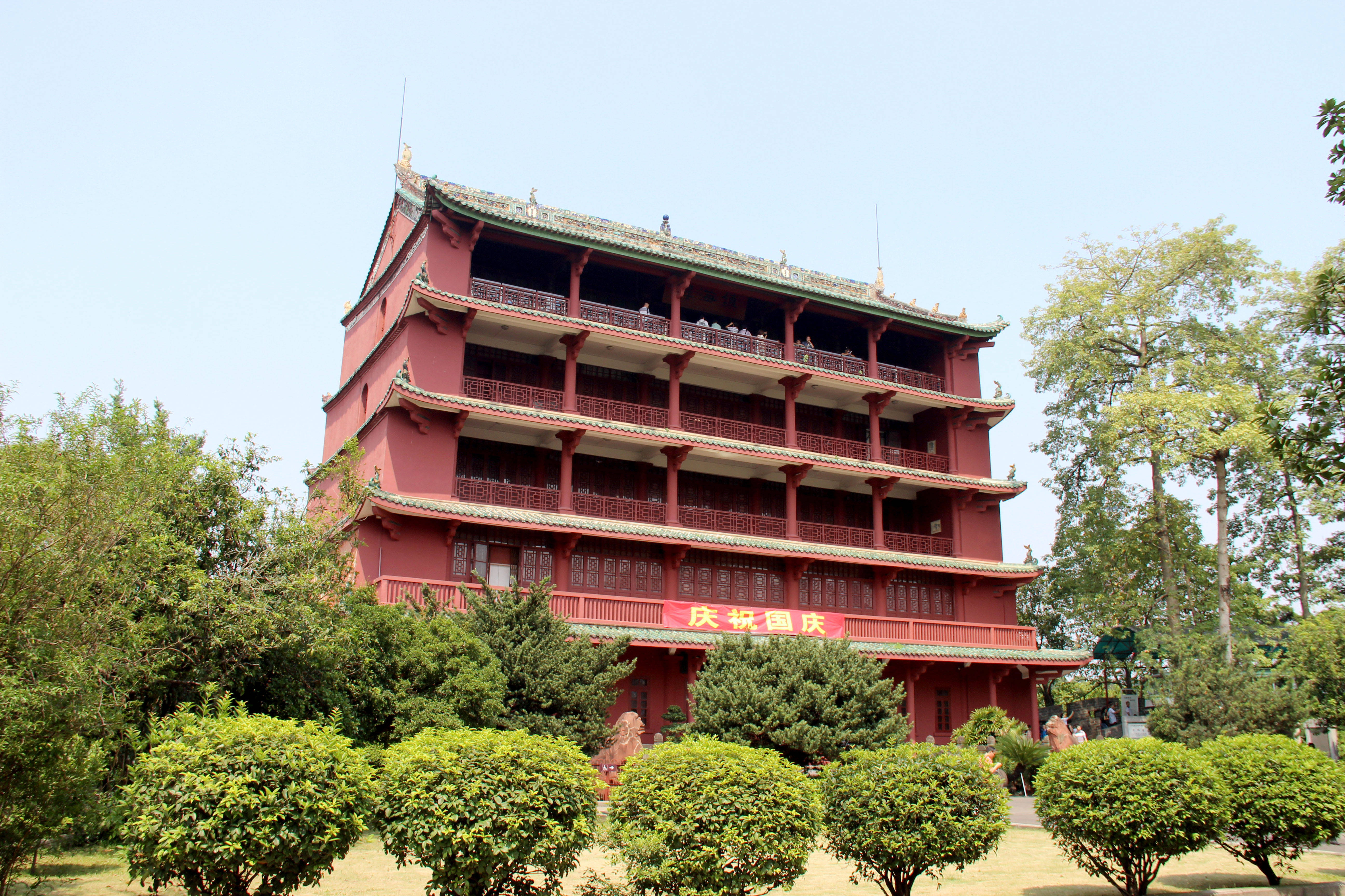 廣州鎮海塔 壁紙 — 一座宏偉的紅色傳統中式寶塔，配以綠色瓦片屋頂，在晴朗的藍天下矗立於茂密的綠樹和修剪整齊的灌木叢中。