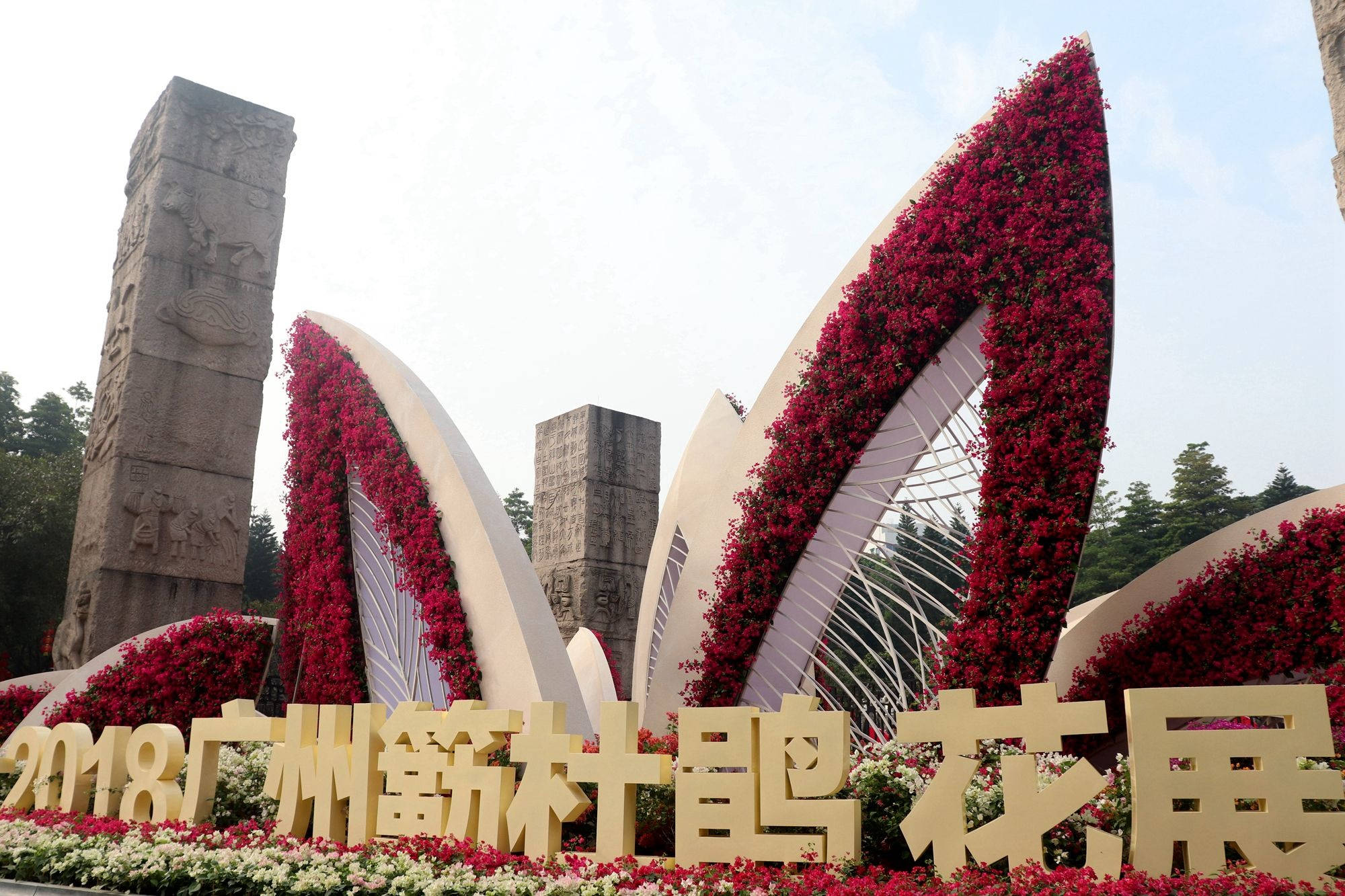 廣州雕塑公園美景 壁紙 — 一個大型花卉裝置，紅色花朵在明亮的天空下沿著白色拱門和石柱傾瀉而下。