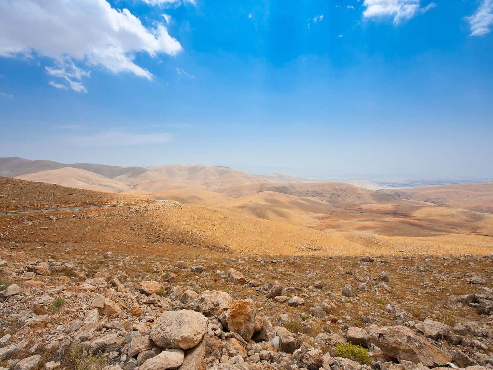 Israel 桌布 壁紙 — 廣闊乾旱的景觀，起伏的金色山丘點綴著岩石，頭頂是明亮的藍天，散佈著雲朵。