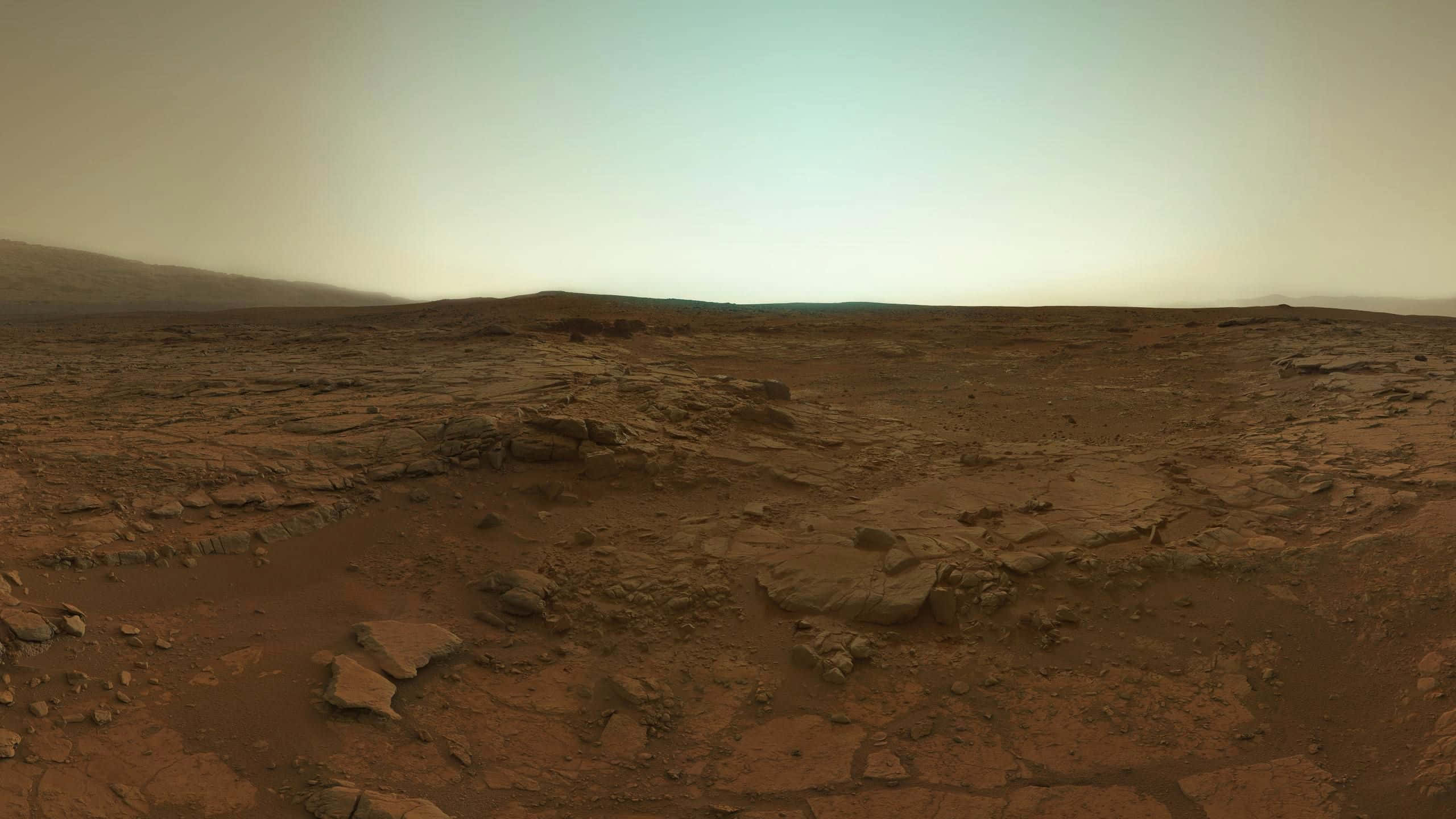 Mars Landscape 桌布 壁紙 — 廣闊嘅鏽紅色火星平原喺朦朧天空下延伸，遠處有起伏嘅山丘同散落嘅岩石。
