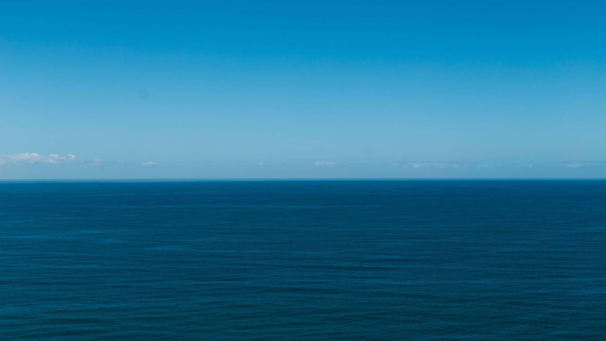 Minimalist Ocean 桌布 壁紙 — 廣闊平靜的海洋與晴朗的藍天在明亮的日光下相遇，地平線上有遙遠的雲朵。