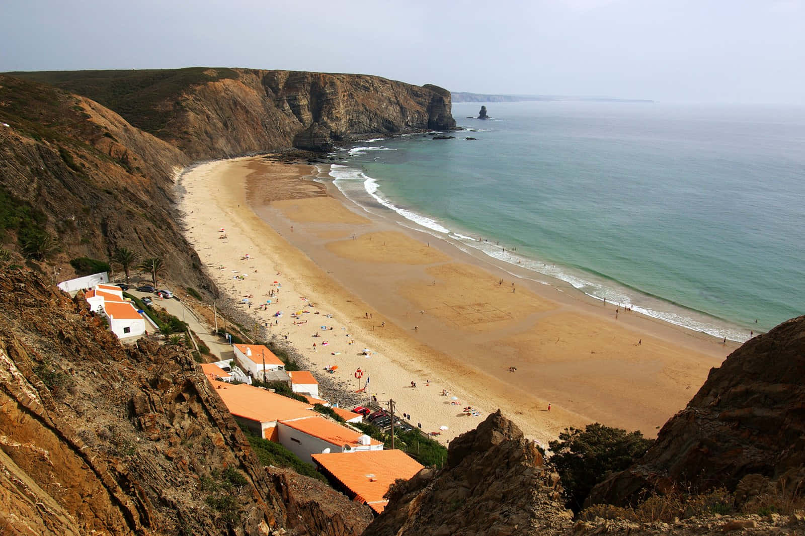 Portuguese Beach 桌布 壁紙 — 廣闊的海岸景觀，沙灘坐落在崎嶇的懸崖之間，橙紅色屋頂的房子緊貼在山坡上。