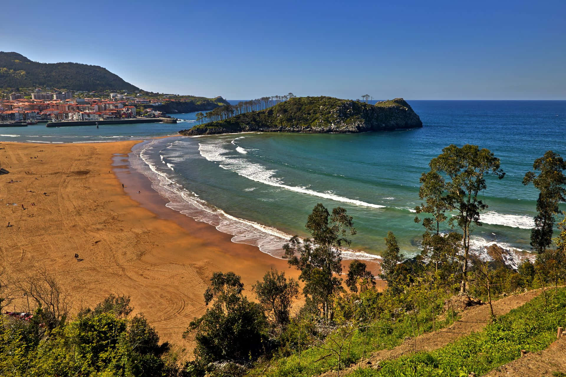 Spanish Beach 桌布 壁紙 — 廣闊的海岸景觀，金色沙灘蜿蜒環繞著碧綠的海水，山坡小鎮和森林覆蓋的懸崖在清澈的藍天下。