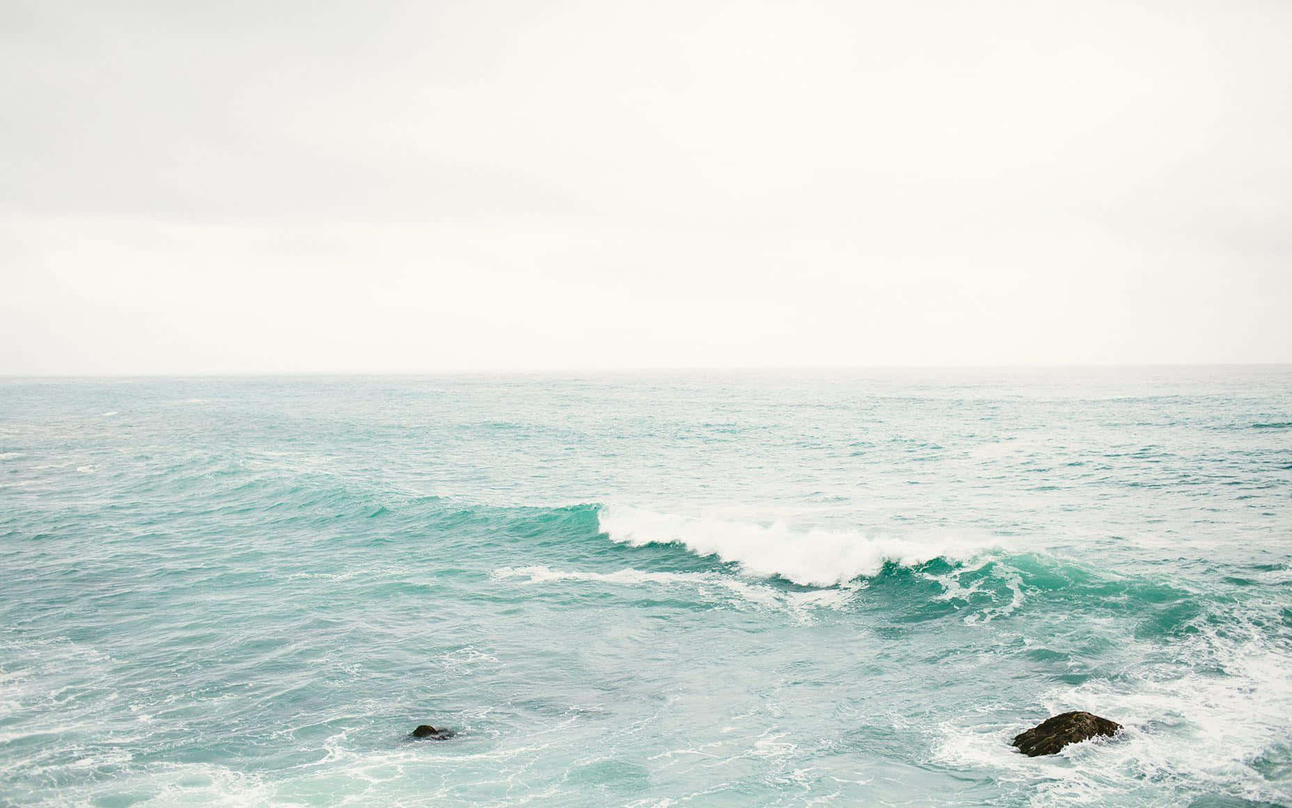 Aesthetic Beach 桌布 壁紙 — 廣闊的海洋，在陰天之下，碧綠色的波浪拍打著深色的岩石。