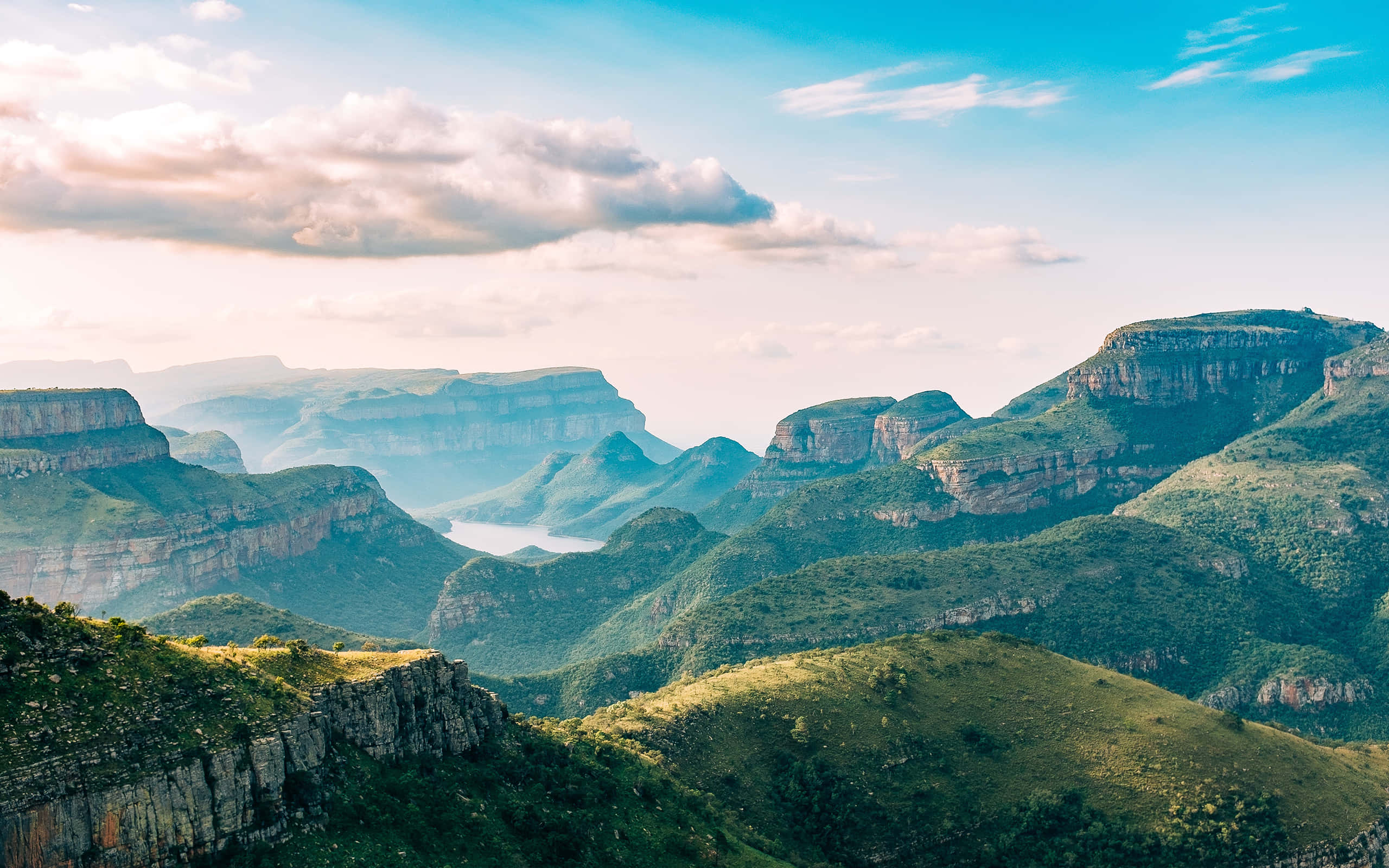 South Africa 桌布 壁紙 — 廣闊的綠色峽谷，擁有層疊的懸崖和遠處的湖泊，在明亮的藍天和柔軟的白雲下。