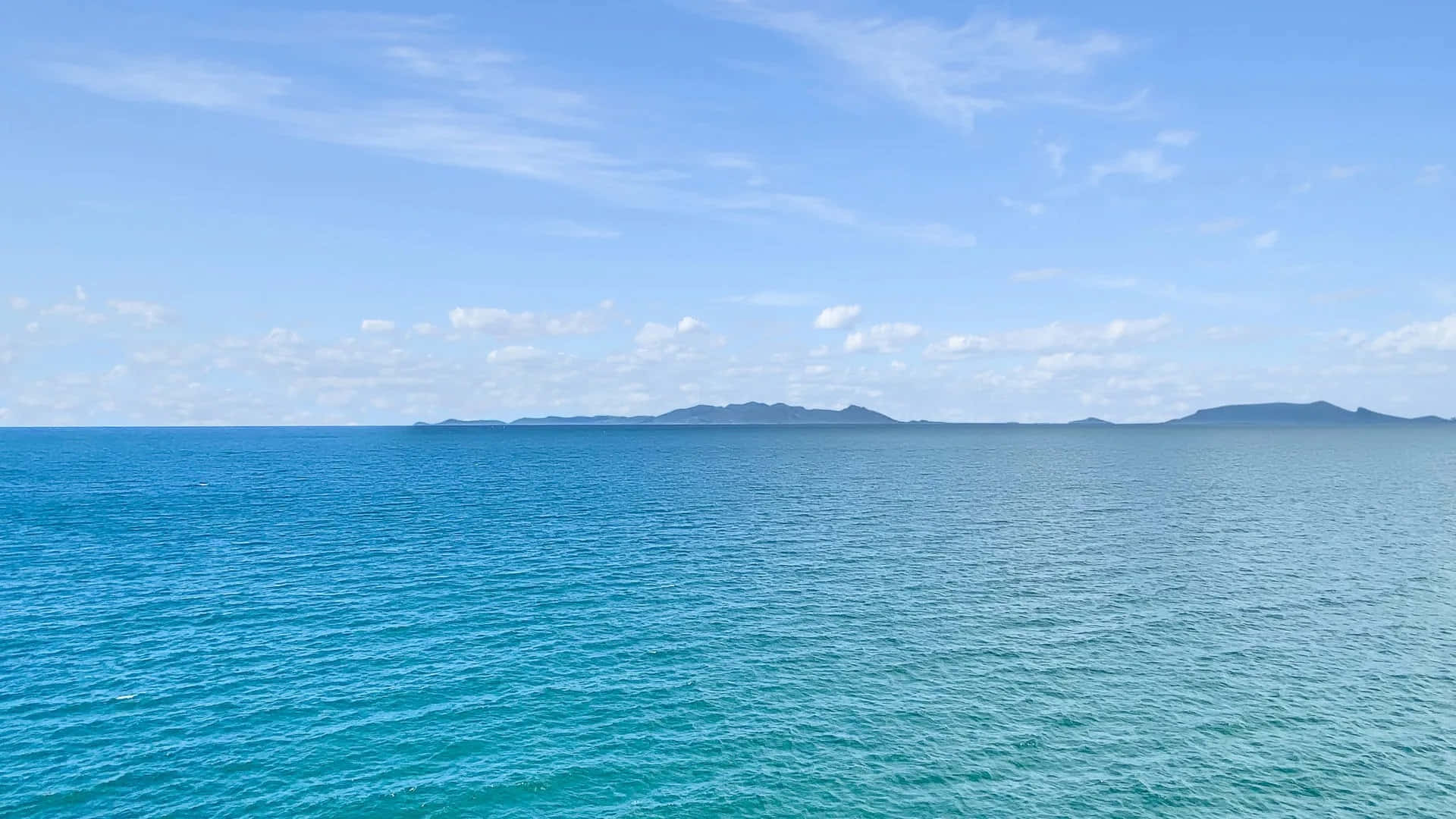 Horizon 桌布 壁紙 — 廣闊的藍綠色海洋延伸至遙遠朦朧的島嶼，天空湛藍，雲朵輕柔。
