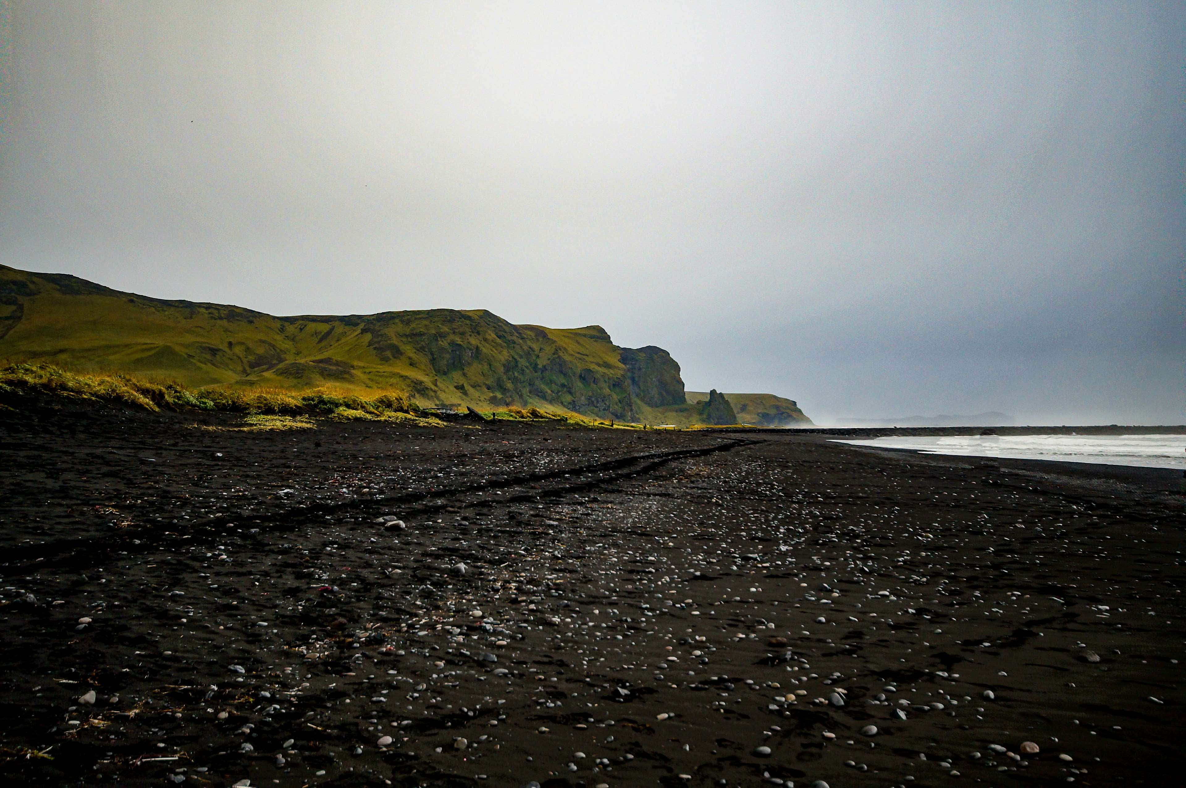 Black Sand Beach 桌布 壁紙 — 廣闊的黑色火山沙灘在陰天下延伸至長滿苔蘚的綠色懸崖。