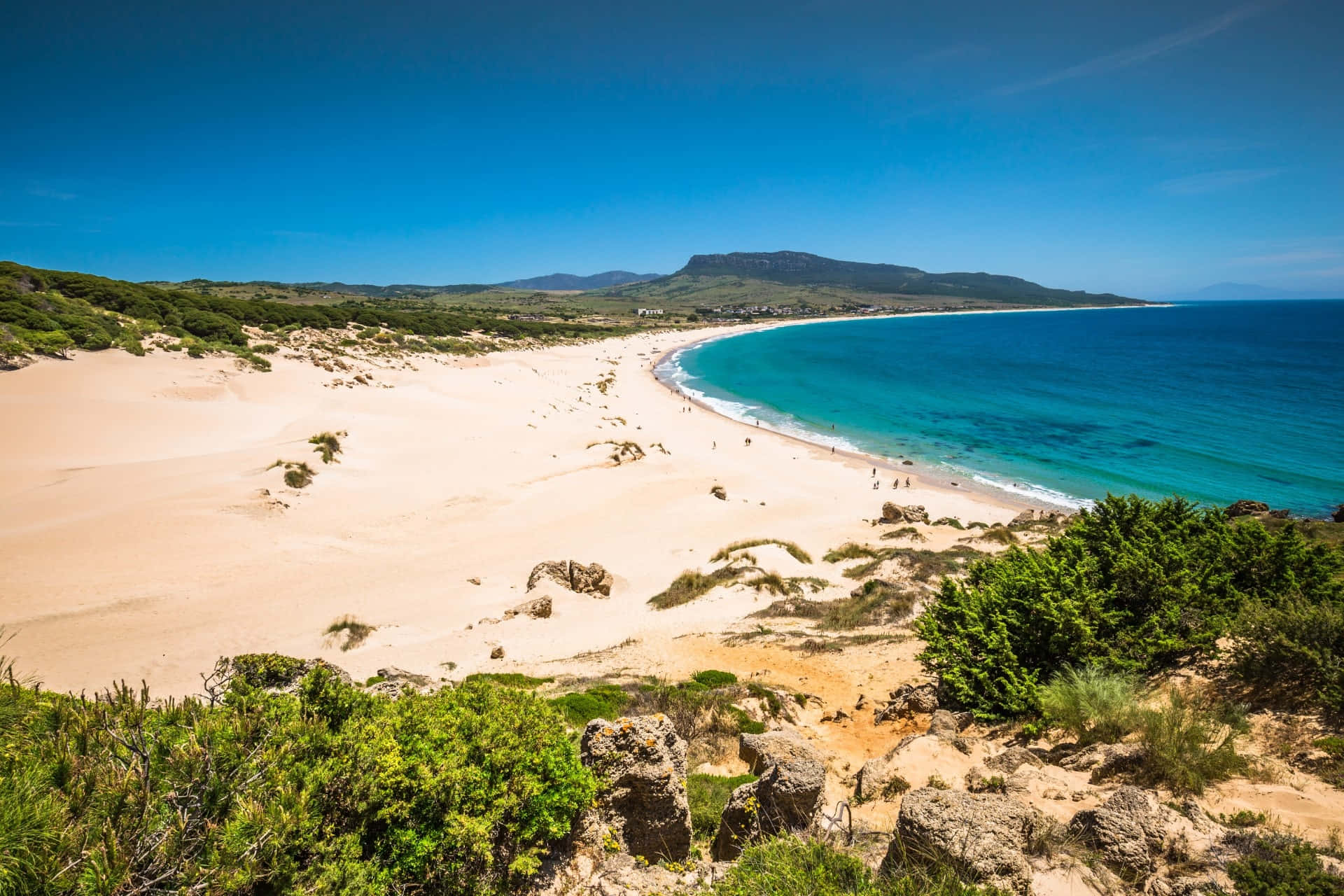 Spanish Beach 桌布 壁紙 — 廣闊視野，彎曲的綠松石色海灘，金色沙灘，兩旁綠色的沙丘和遠處的山丘，在清澈的藍天下。