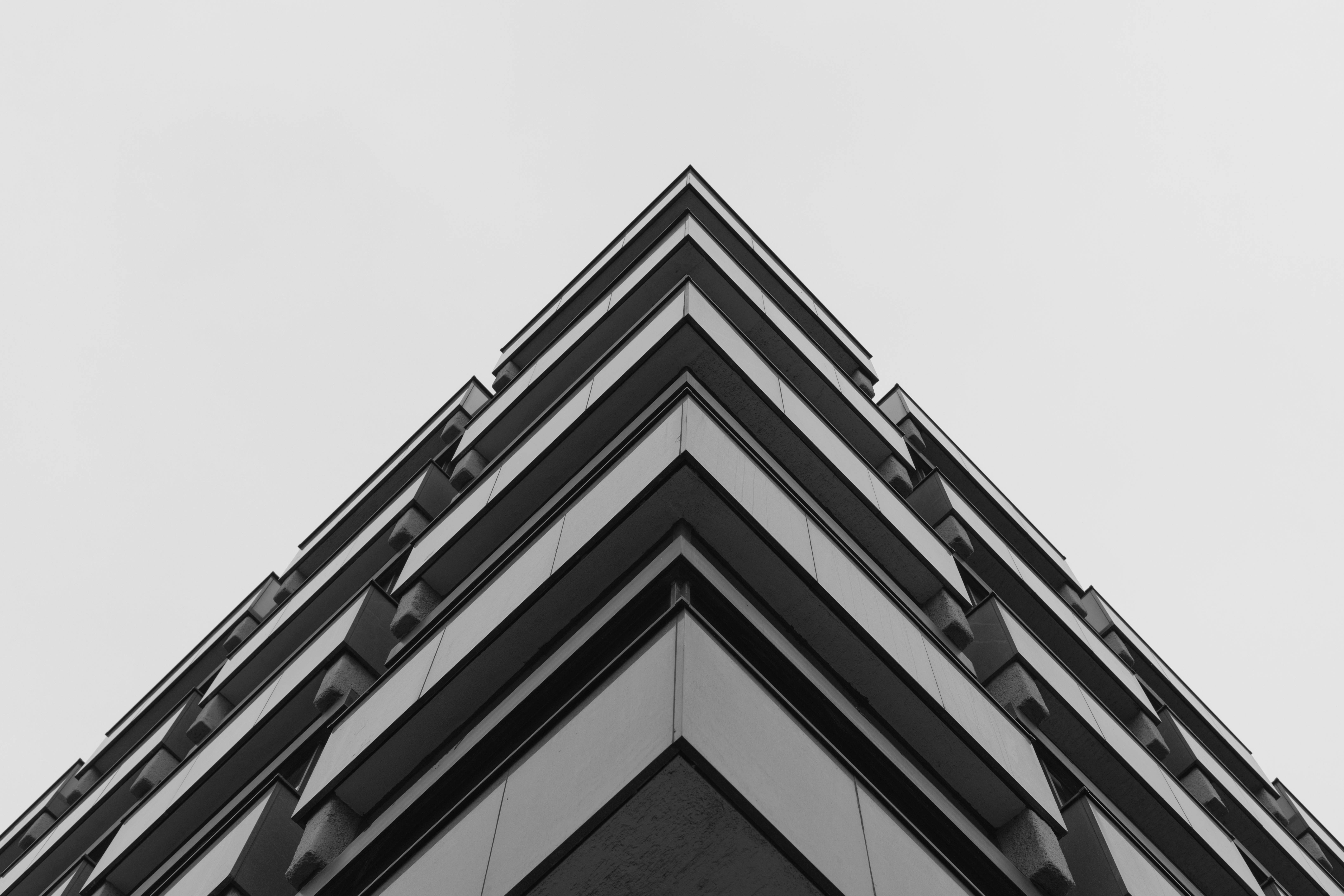 建築卓越之美——鋭邊混凝土大樓 壁紙 — 一張以普通天空為背景，低角度拍攝的現代建築角落的清晰黑白照片。