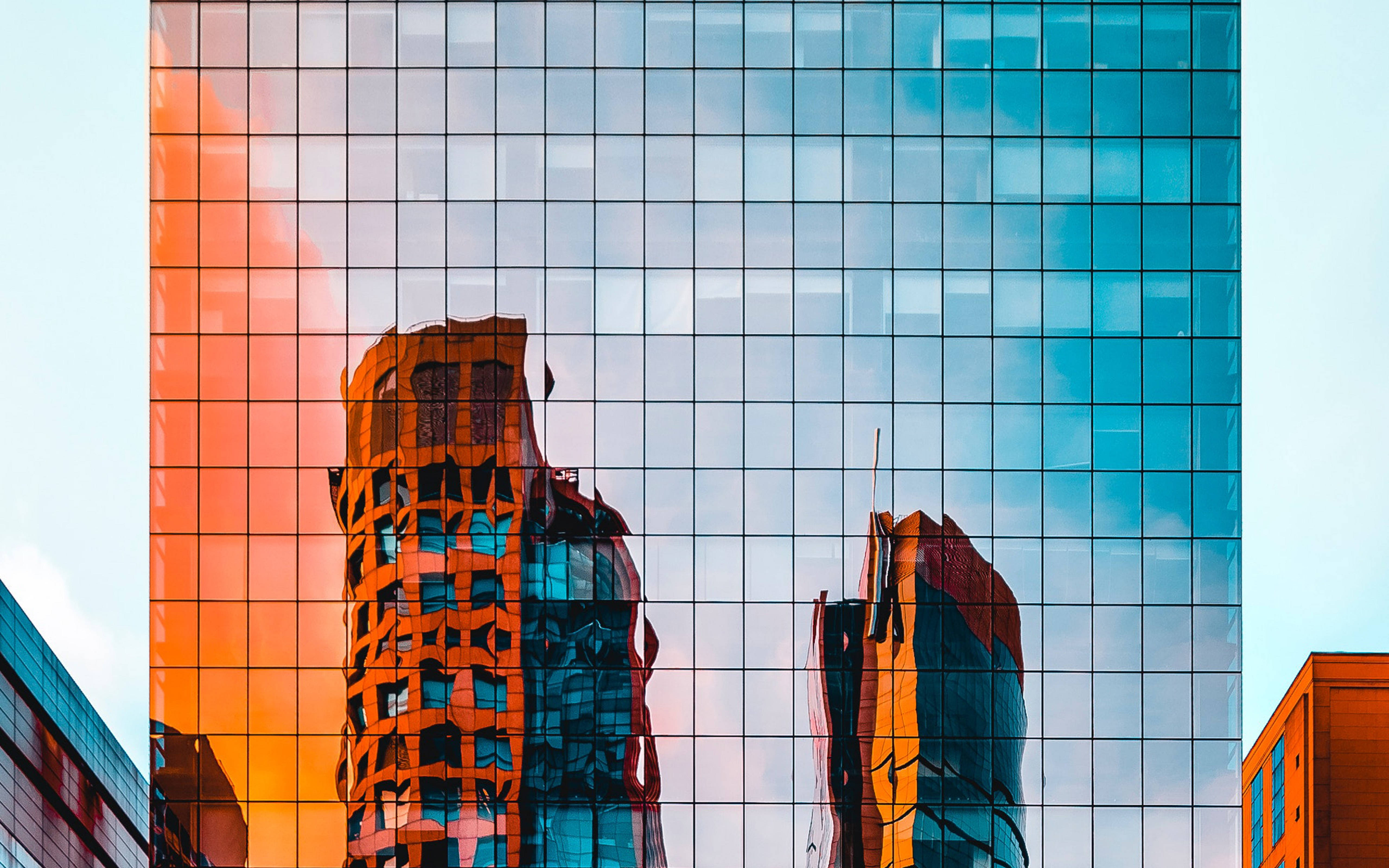 建築外牆抽象倒影 壁紙 — 一座玻璃摩天大樓在蒼白的天空映襯下，反射出火橙色和冷藍色的摩天大樓。