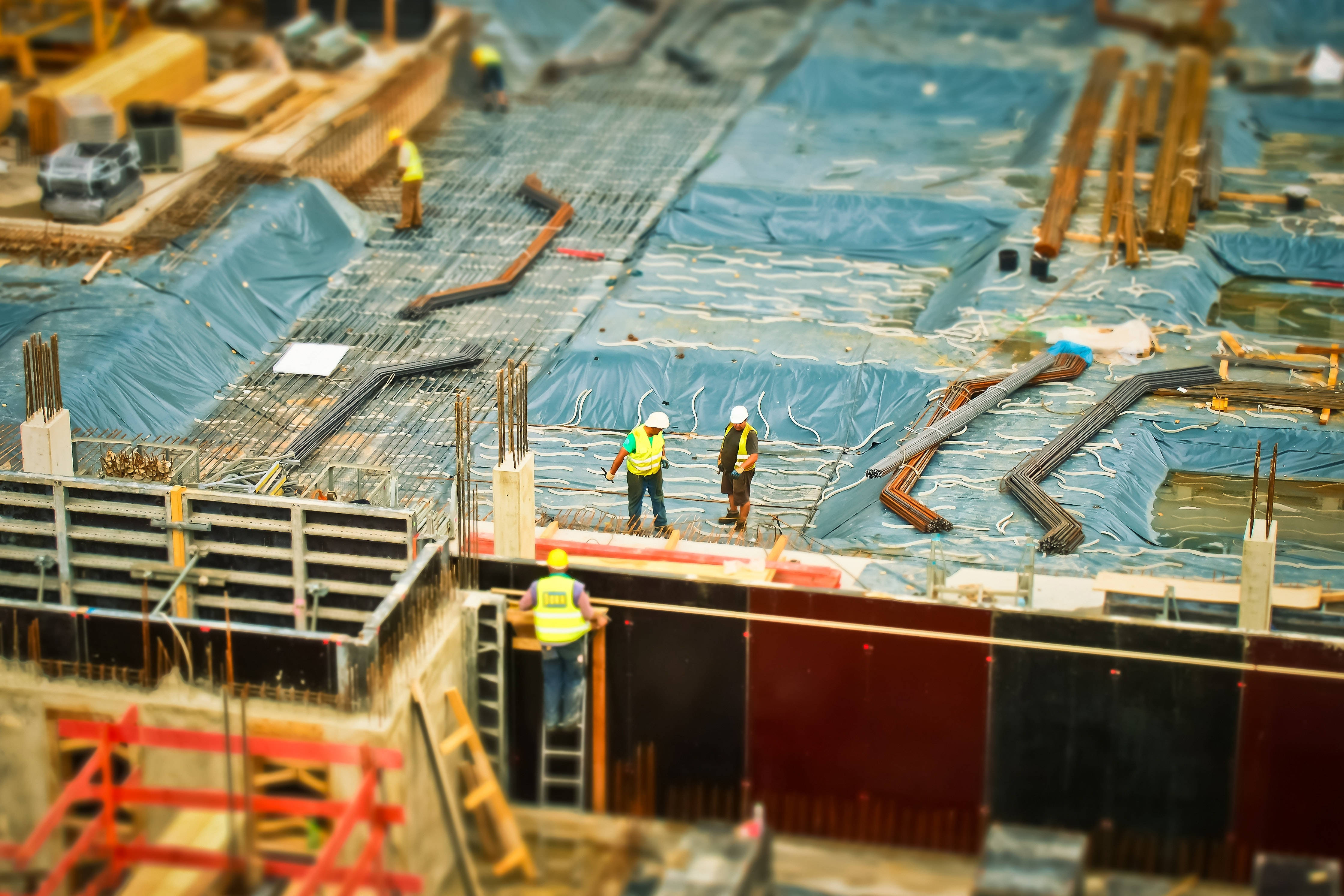 建築工人在工地辛勤作業 壁紙 — 細小嘅建築工人著住鮮色背心，企喺一個鋪咗藍色防水布、有鋼筋同棚架嘅廣闊工地上面，喺溫暖嘅日光下。