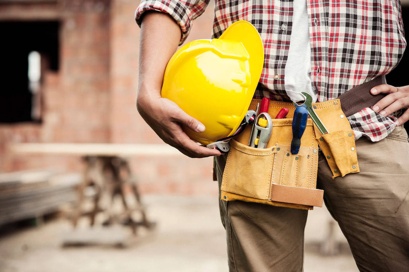 建築工人腰間掛著工具 壁紙 — 一名建築工人在磚砌工地拿著亮黃色安全帽，同時佩戴裝滿各種工具的工具腰帶。