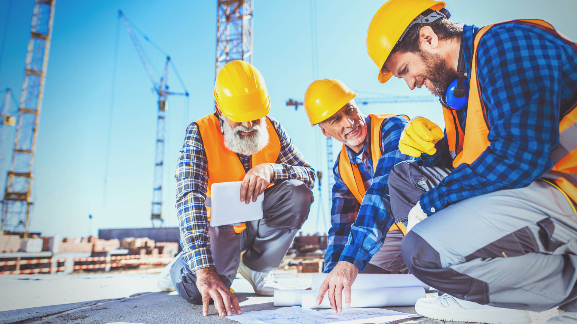 建築工人與工程師 壁紙 — 三名戴著安全帽和背心的微笑建築工人在陽光明媚的建築工地上審查藍圖，背景有起重機。