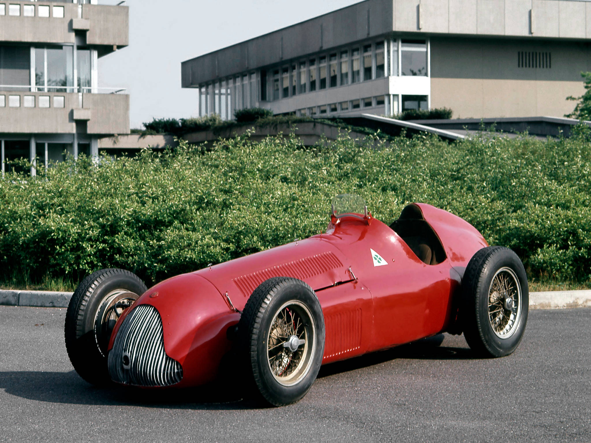 建築後方停車場紅色Alfa Romeo iPhone壁紙 壁紙 — 一輛光澤的紅色復古賽車配備鋼絲輪，停在瀝青路面上，前方有綠色灌木和現代建築，在明亮的日光下。