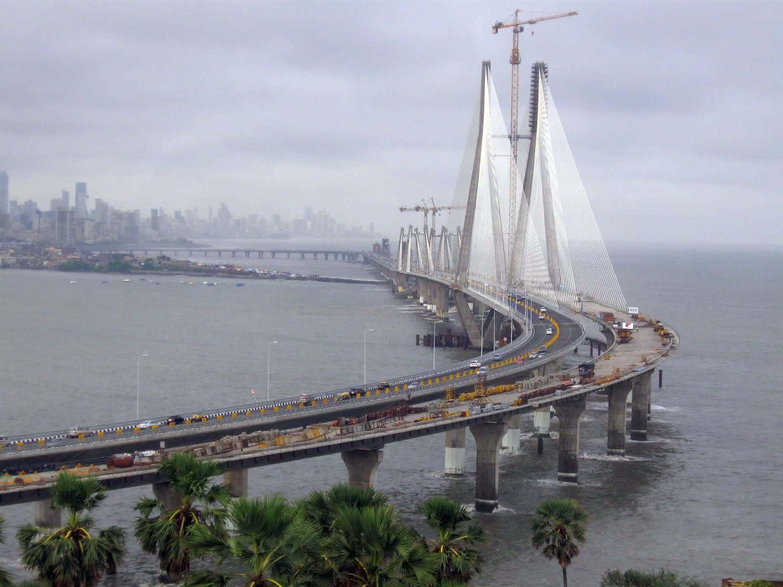 Mumbai 桌布 壁紙 — 建設中的薩哈卡爾尼大橋的廣闊景觀，起重機高聳於其纜索之上，纜索橫跨寬闊的海灣，伸向朦朧的城市天際線。