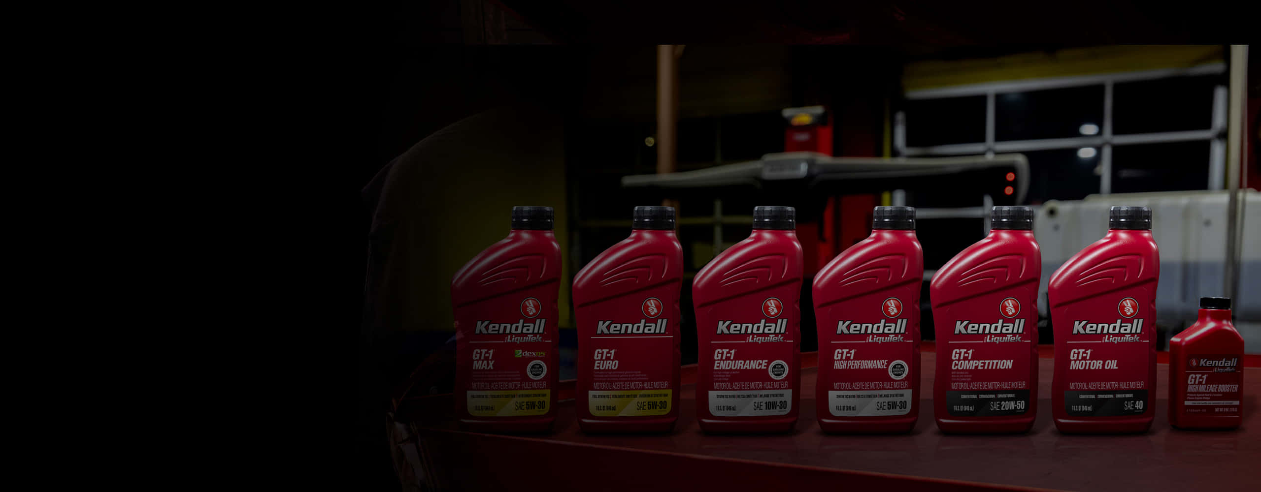 引擎維護的必要元素 壁紙 — 一排紅色Kendall機油瓶整齊排列在昏暗車庫的架子上，背景中有一位技工。