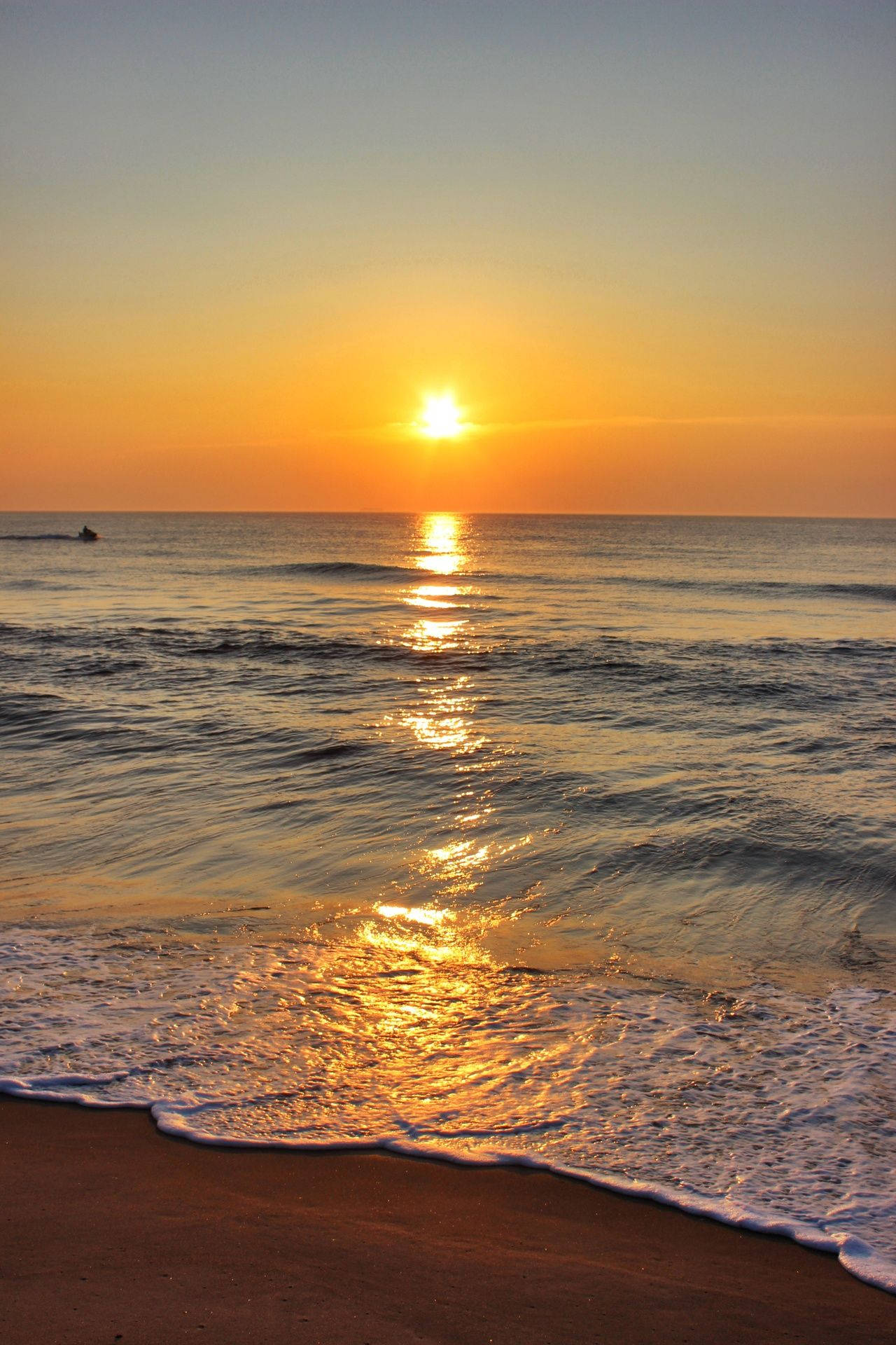 弗吉尼亞海灘橙藍天空日落 壁紙 — 金色夕陽照耀在平靜的海浪上，沖刷著沙灘，遠處有游泳者。
