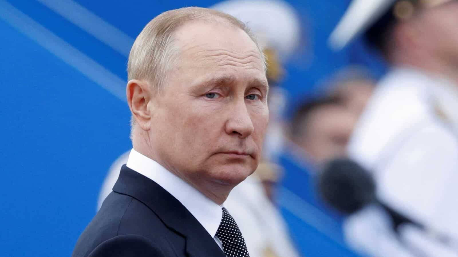 弗拉基米尔·普京側目凝視 壁紙 — 弗拉基米爾·普京身穿深色西裝打領帶的特寫肖像，背景為鮮豔的藍色，帶有模糊的人物。