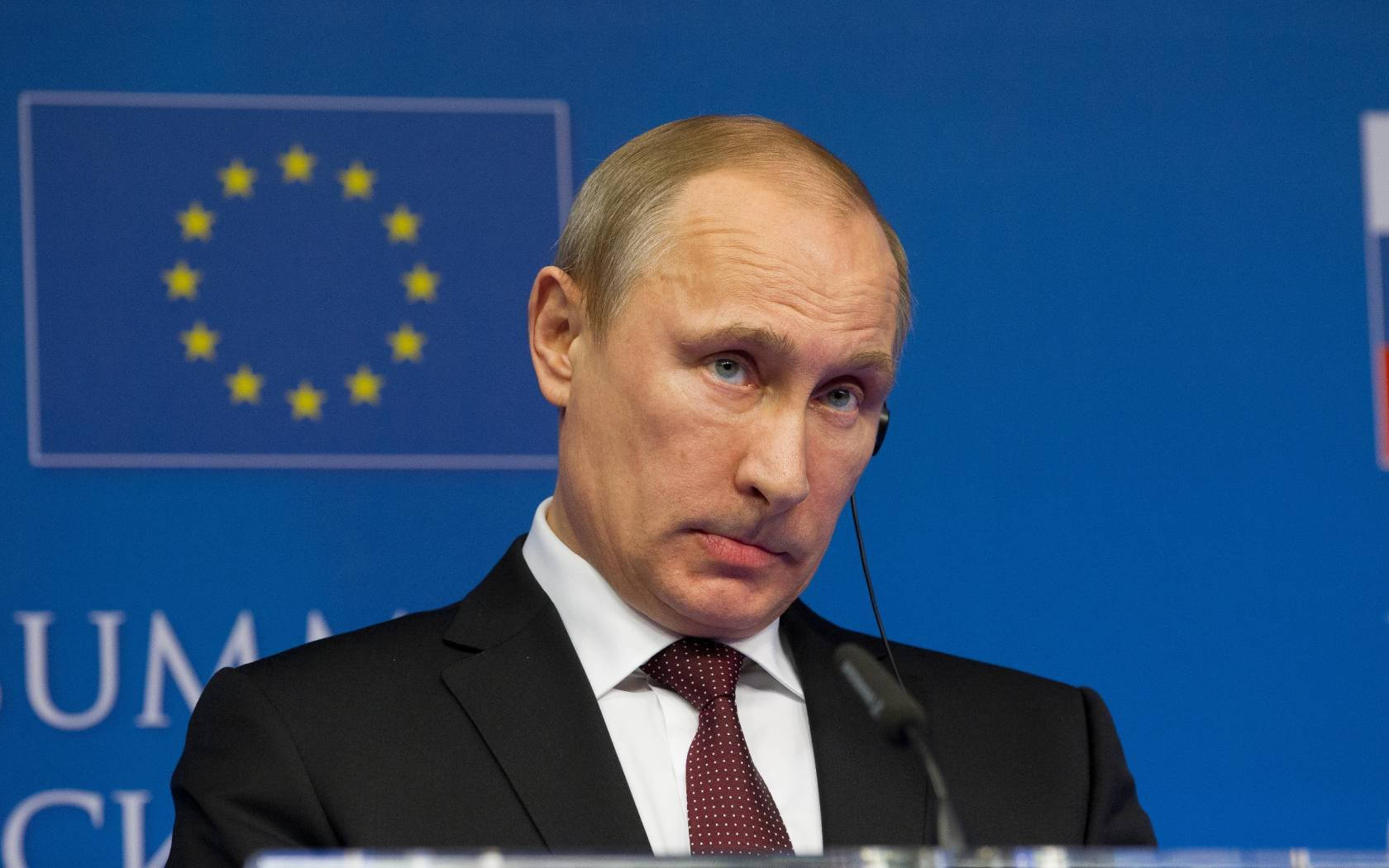弗拉基米尔·普京嘴唇微噘 壁紙 — Vladimir Putin 身穿深色西裝和紅色領帶，在背後有藍色歐盟旗幟的講台上發言。