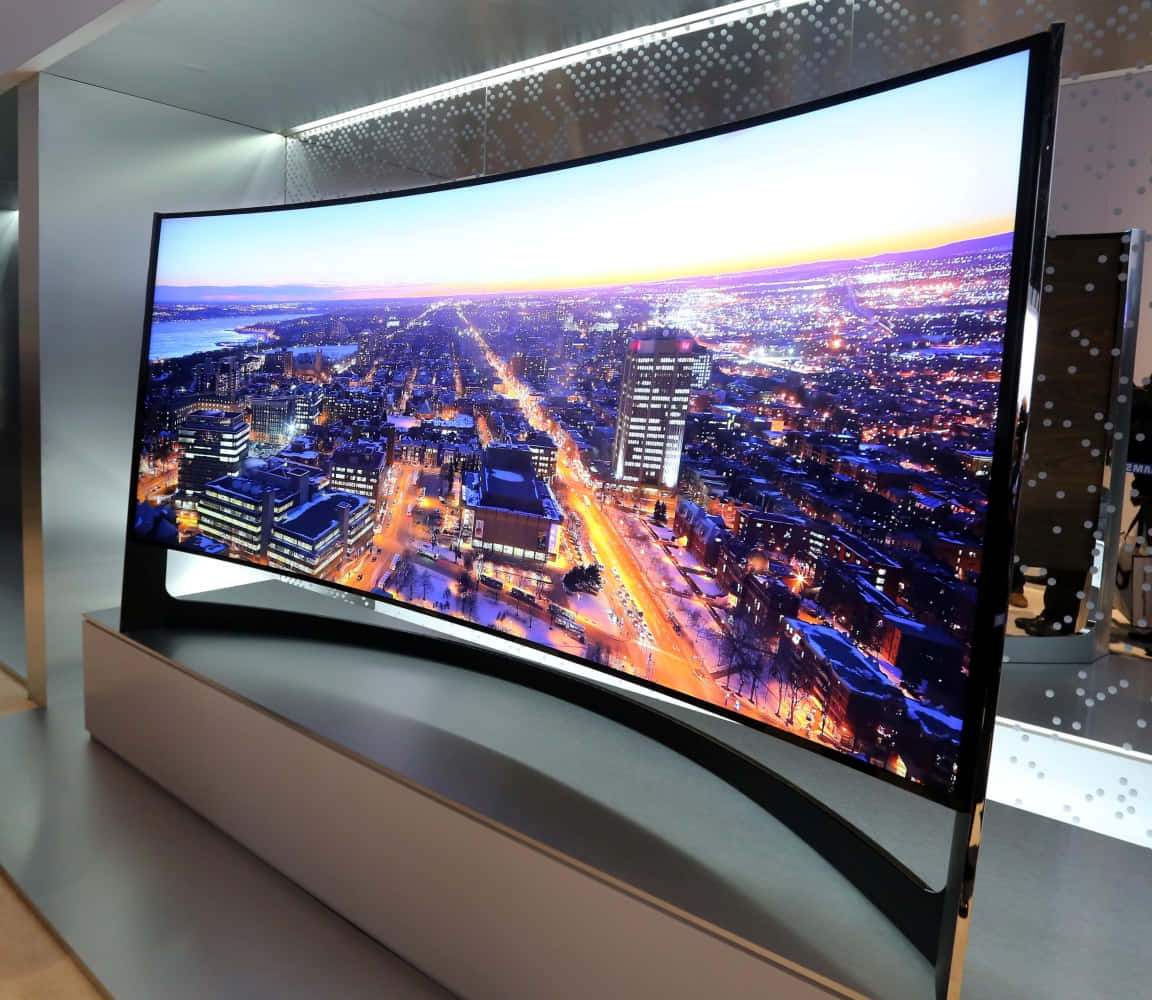 tv 桌布 壁紙 — 弧形超寬電視顯示出暮色中充滿活力的雪景城市風光，有發光的街燈和明亮的建築物。