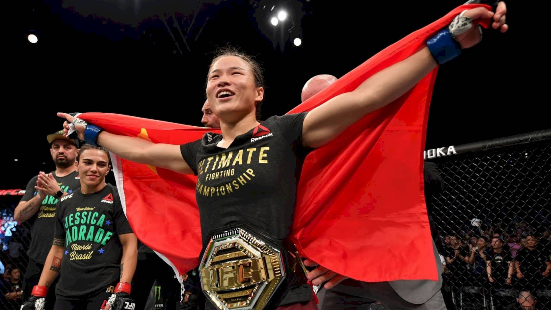 女性MMA拳手在八角籠中手持冠軍腰帶和紅旗慶祝勝利。
