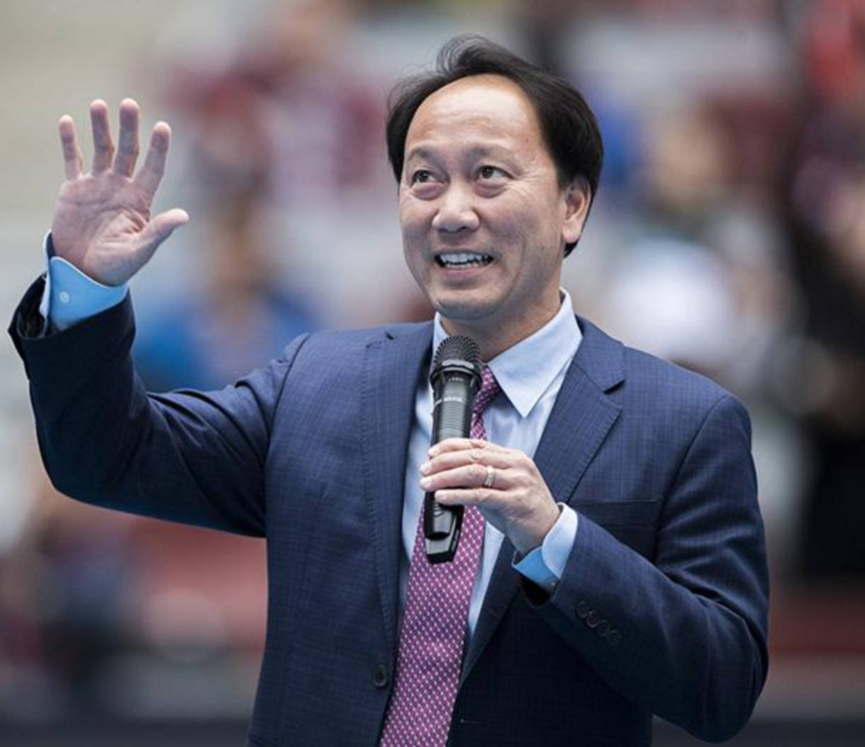 張德培演講 壁紙 — 一個微笑的西裝男子在模糊的人群前揮手，同時拿著麥克風。
