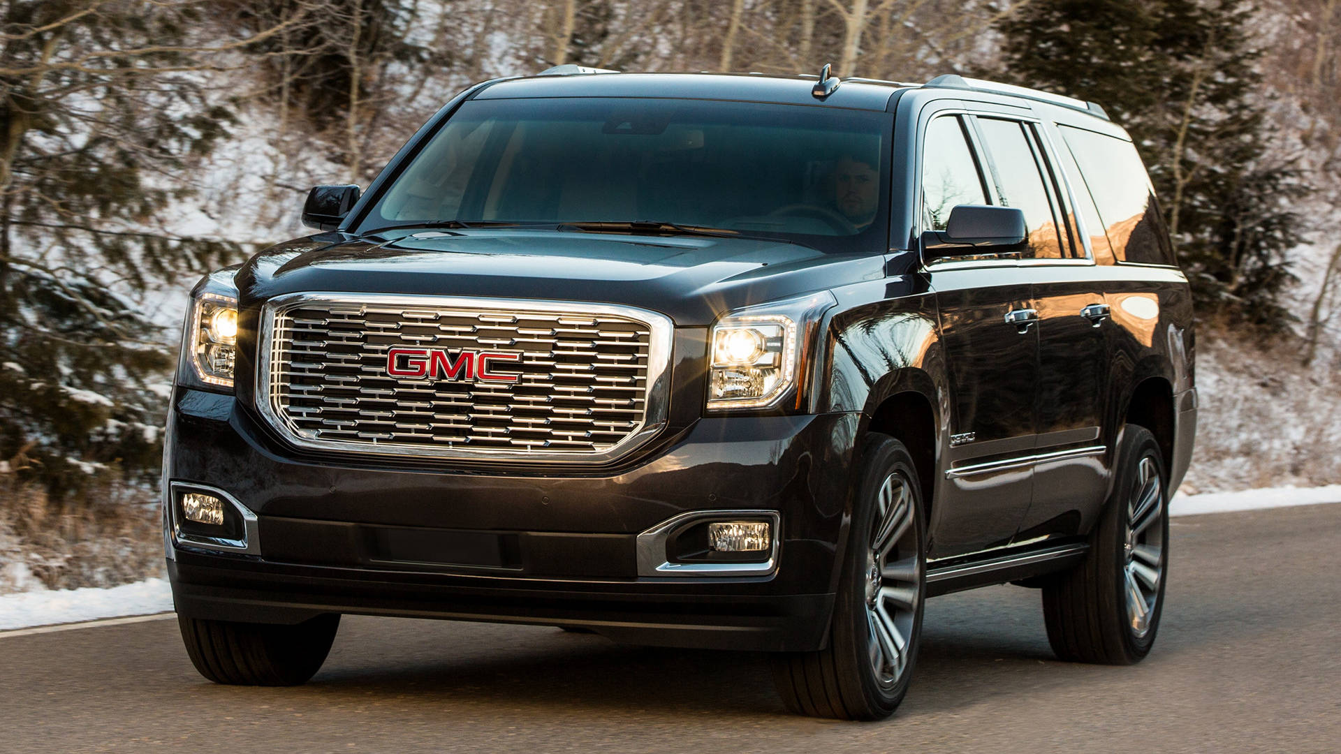 強勁 GMC Yukon 行駛於開放道路 壁紙 — 一輛時尚的黑色GMC Yukon SUV在柔和的日光下行駛在一條被冬日禿樹環繞的雪路上。