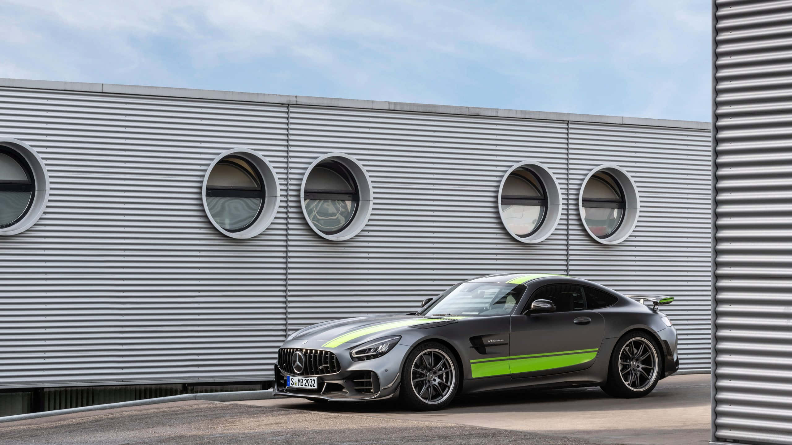 強勁優雅 - AMG GT R 背景 壁紙 — 一輛時尚的灰色 Mercedes-AMG GT 帶有螢光綠色點綴，停在一個帶有圓形窗戶的現代波紋金屬建築前。