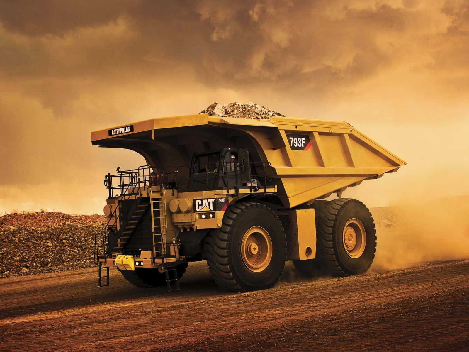 強勁堅固的黃色自卸車正運載貨物 壁紙 — 一輛巨大的黃色Caterpillar 793F礦用自卸車在金色天空下，穿越塵土飛揚的露天礦場運送礫石。