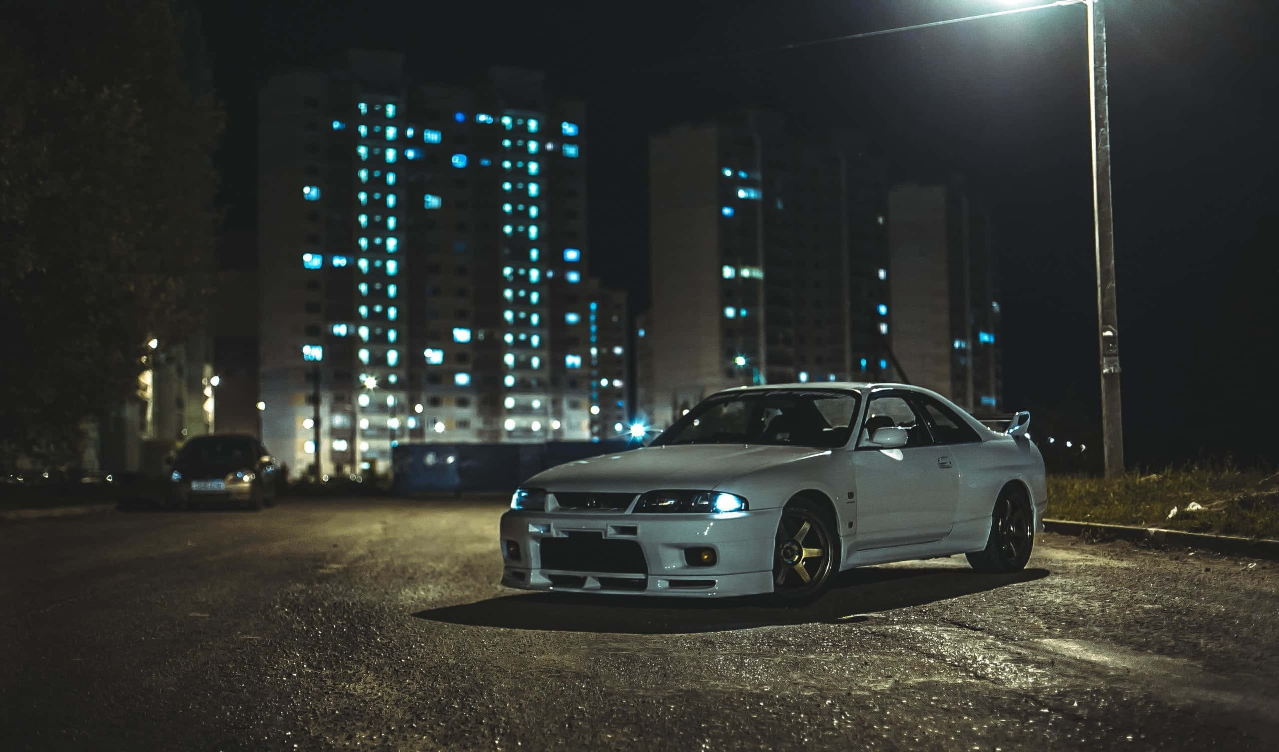 強勁增壓：Nissan R33 GTR 壁紙 — 一輛白色跑車在夜間濕潤的城市街道上，在街燈下閃閃發光，背景是發光的公寓大樓。