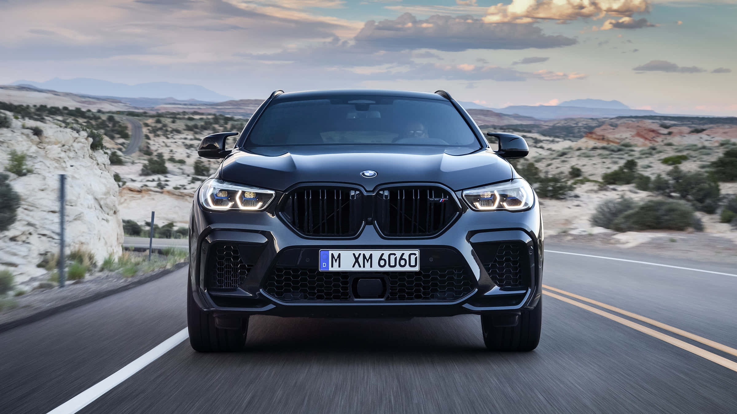 強勁的 BMW X6 M 壁紙 — 一輛時尚的黑色BMW XM 6060 SUV在沙漠公路上向前行駛，天空多雲，遠處有山脈。