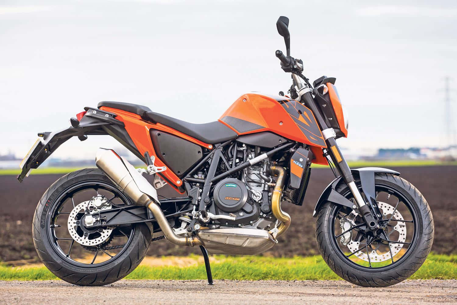 強勁的KTM Duke摩托車 壁紙 — 一架搶眼的橙色和黑色KTM電單車，在陰天之下，停泊在犁過的田地旁邊的碎石路上。