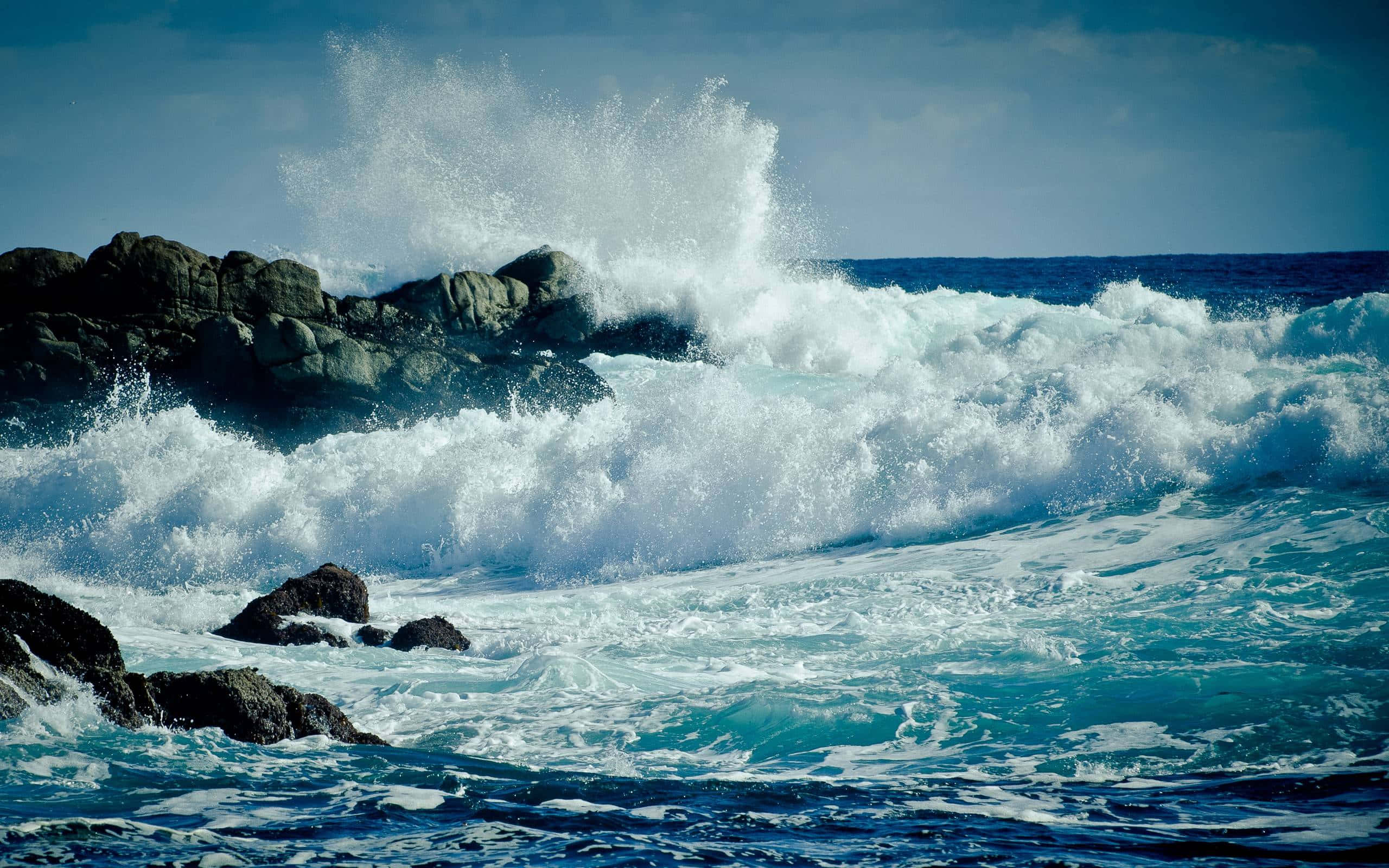 waves 桌布 壁紙 — 強勁的海浪拍打著黑暗崎嶇的岩石，在晴朗的藍天下將白色的浪花高高濺起。
