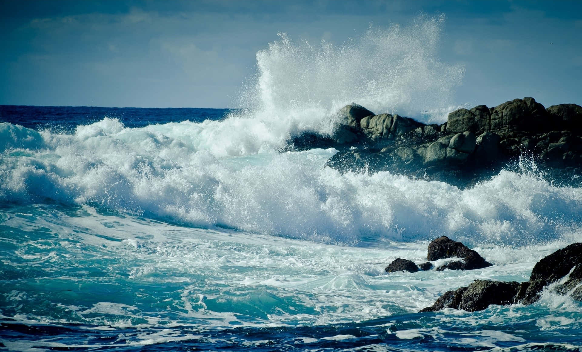 waves 桌布 壁紙 — 強勁的藍綠色波浪拍打著黑暗的鋸齒狀岩石，在晴朗的藍天下將白色浪花高高噴向空中。