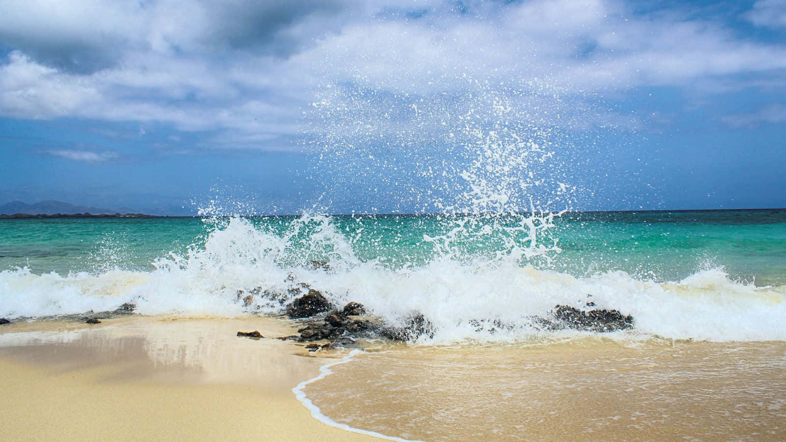 Spanish Beach 桌布 壁紙 — 強勁的藍綠色海浪拍打著金色沙灘上的深色岩石，天空部分多雲，呈現藍色。