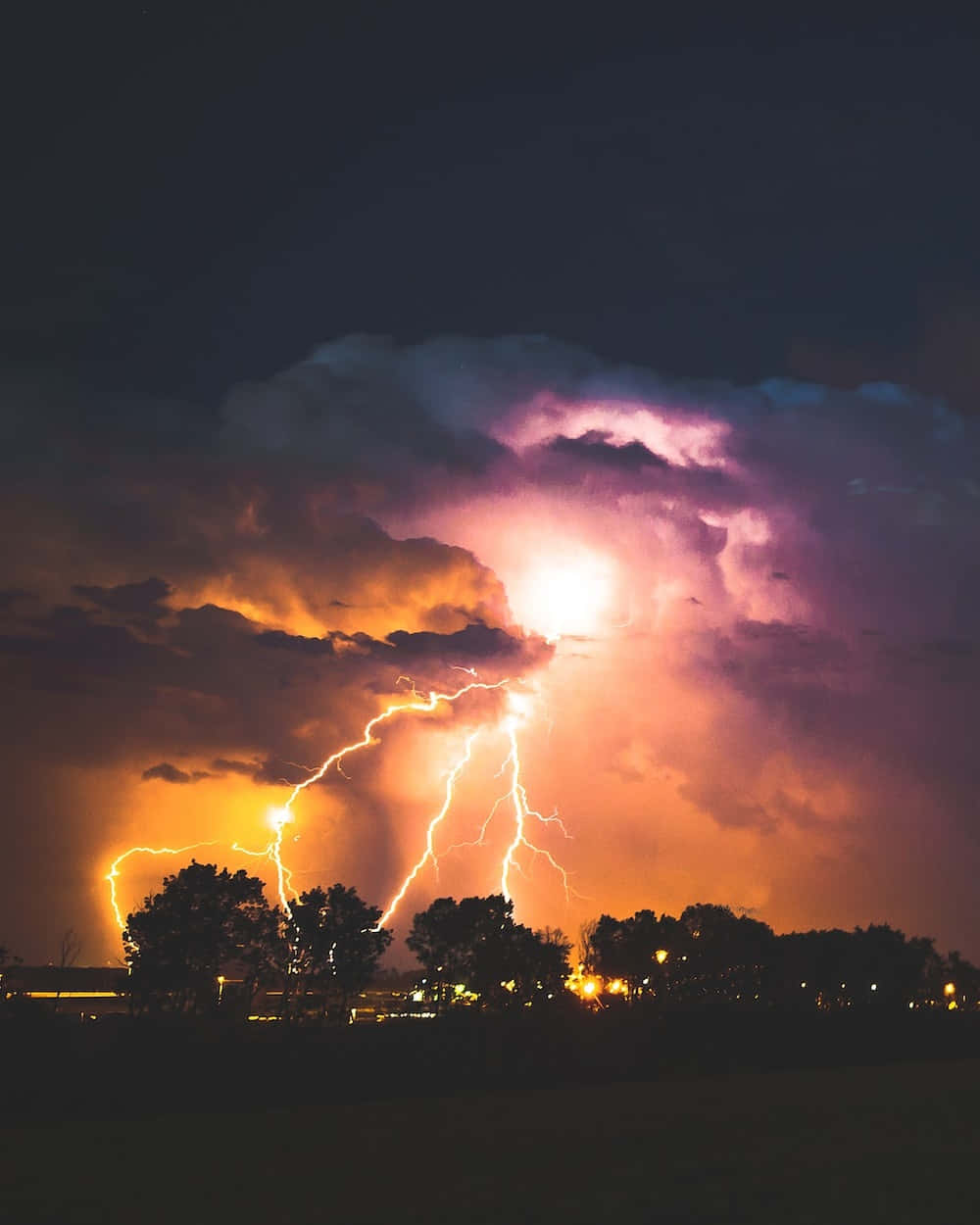 強勁閃電照亮黑暗天空 壁紙 — 一道強勁的閃電照亮了暴風雨的夜空，紫色和橙色的雲層籠罩在剪影樹木和遠處燈光之上。