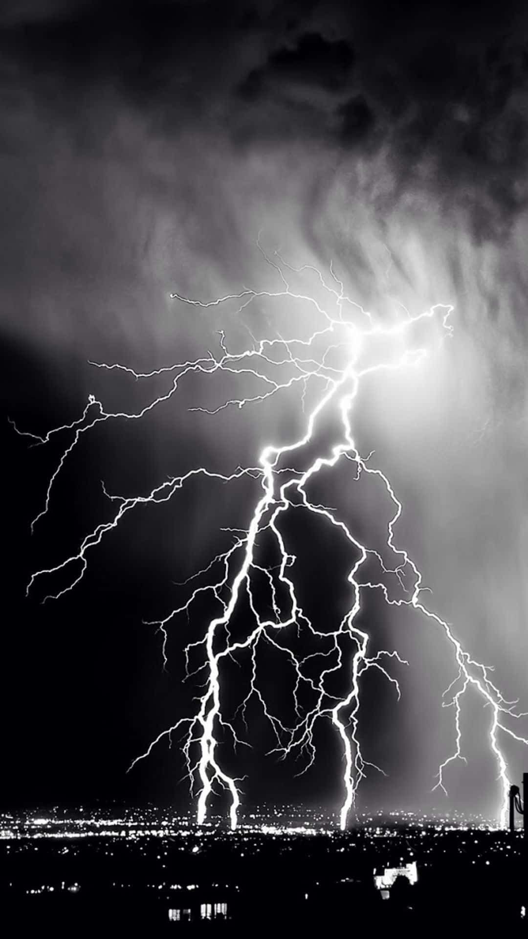 強勁黑白風暴 壁紙 — 一道巨大的閃電在暴風雨的夜空中分叉，照亮了遠處燈火輝煌的城市景觀。