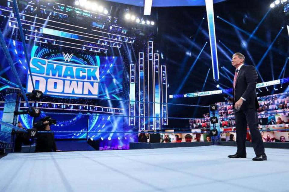 強大的摔角手在擂台中對決 壁紙 — 一位身穿西裝的WWE SmackDown主持人站在摔角擂台邊緣，背後有明亮的藍色舞台燈光和大屏幕。