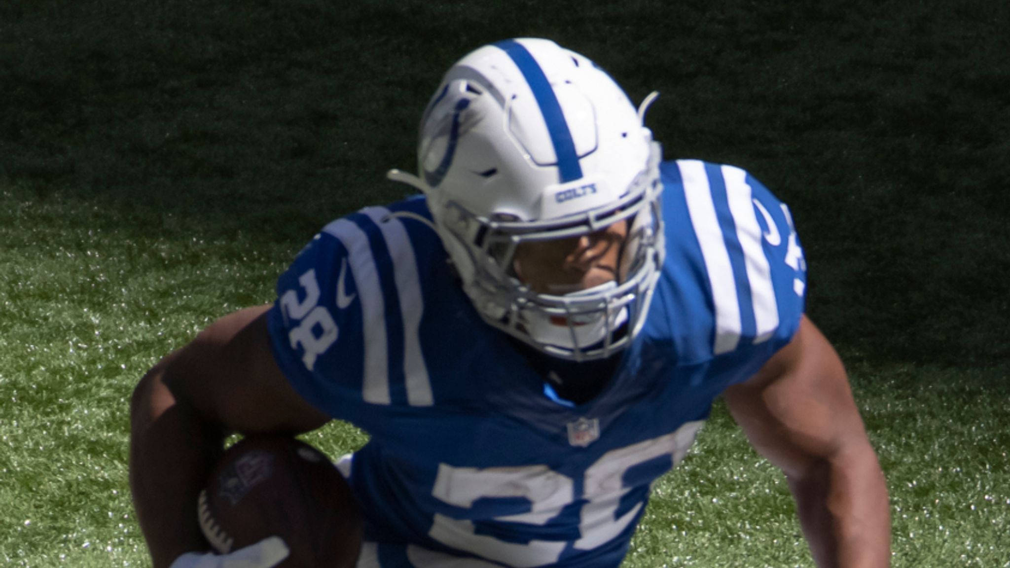 強尼·泰勒頭盔遮眼 壁紙 — 一名身穿藍白 Colts 裝備的美式足球員在陽光明媚的綠色球場上持球衝刺。