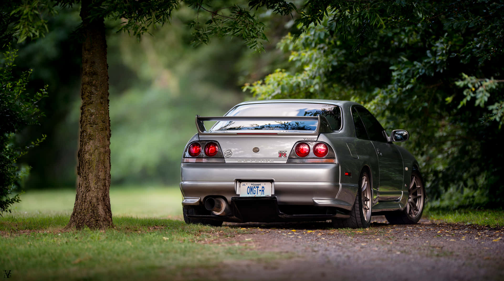 強悍的 Skyline — R33 GTR 壁紙 — 一輛銀色日產Skyline R34停泊在碎石路上，周圍是茂密的綠樹，光線柔和自然。